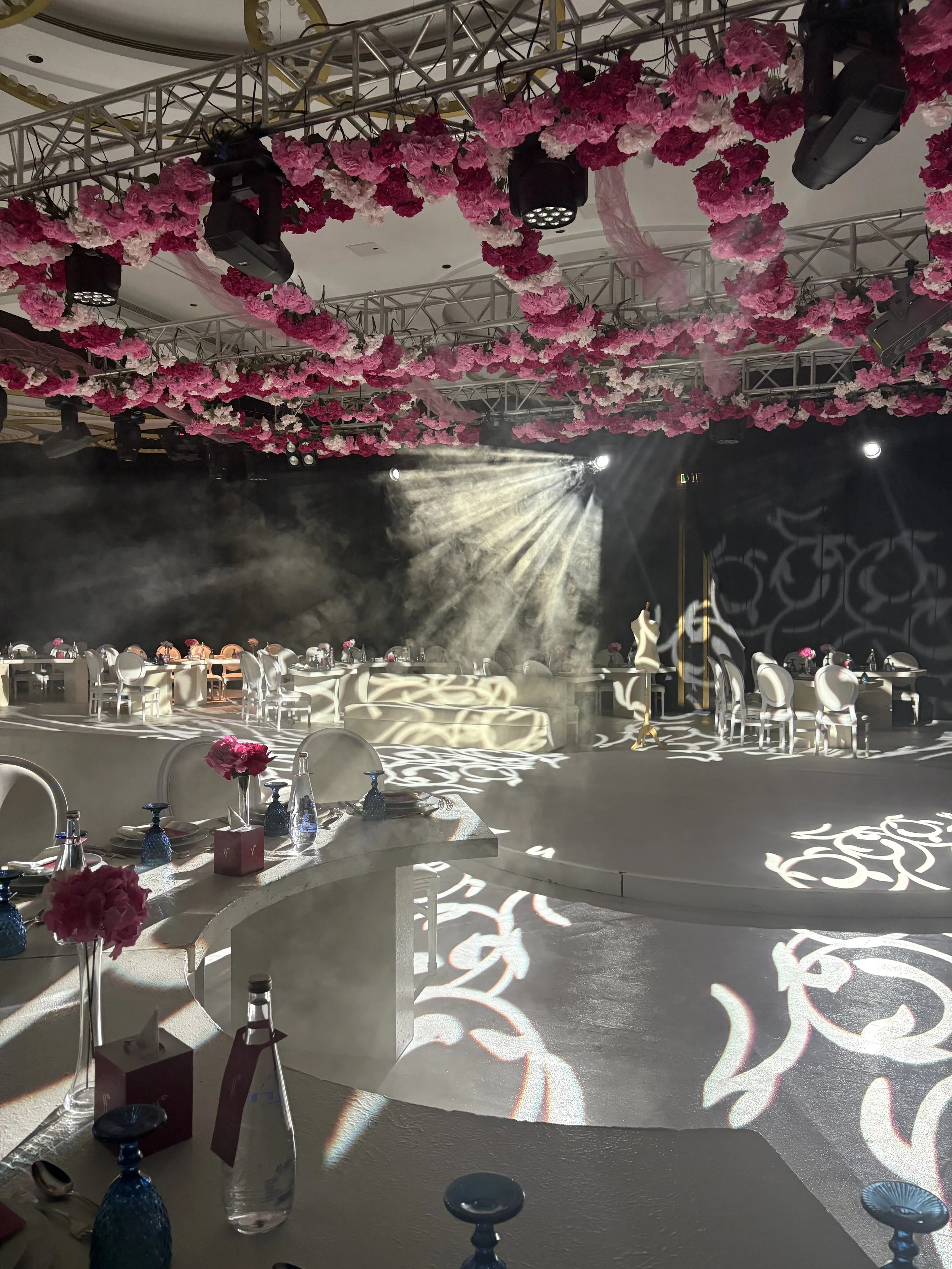 Event space with tables decorated for a celebration, adorned with pink floral arrangements, colorful glassware, and eye-catching light projections on the floors and walls, with pink flowers hanging from the ceiling.