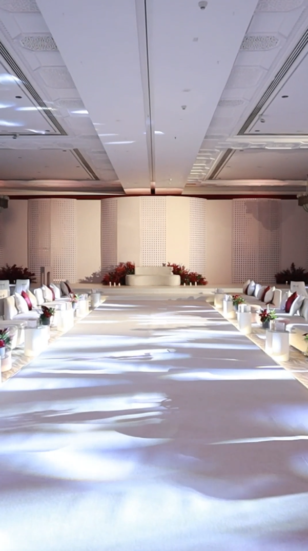 A large, elegant indoor wedding ceremony space with a long white aisle carpet, surrounded by white cushioned chairs with pillows and flowers, leading to a decorated wedding altar with red floral arrangements. Immersive Professional Focused Stage Ligh