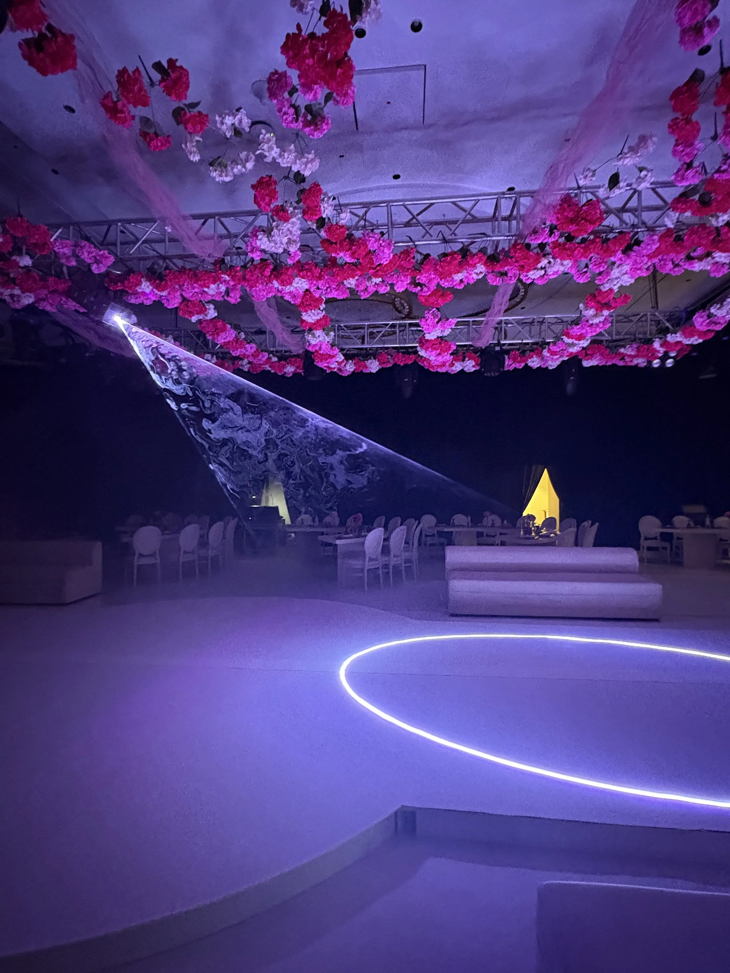 Empty event space with hanging pink and white flower decorations, purple lighting, and modern seating arrangements.