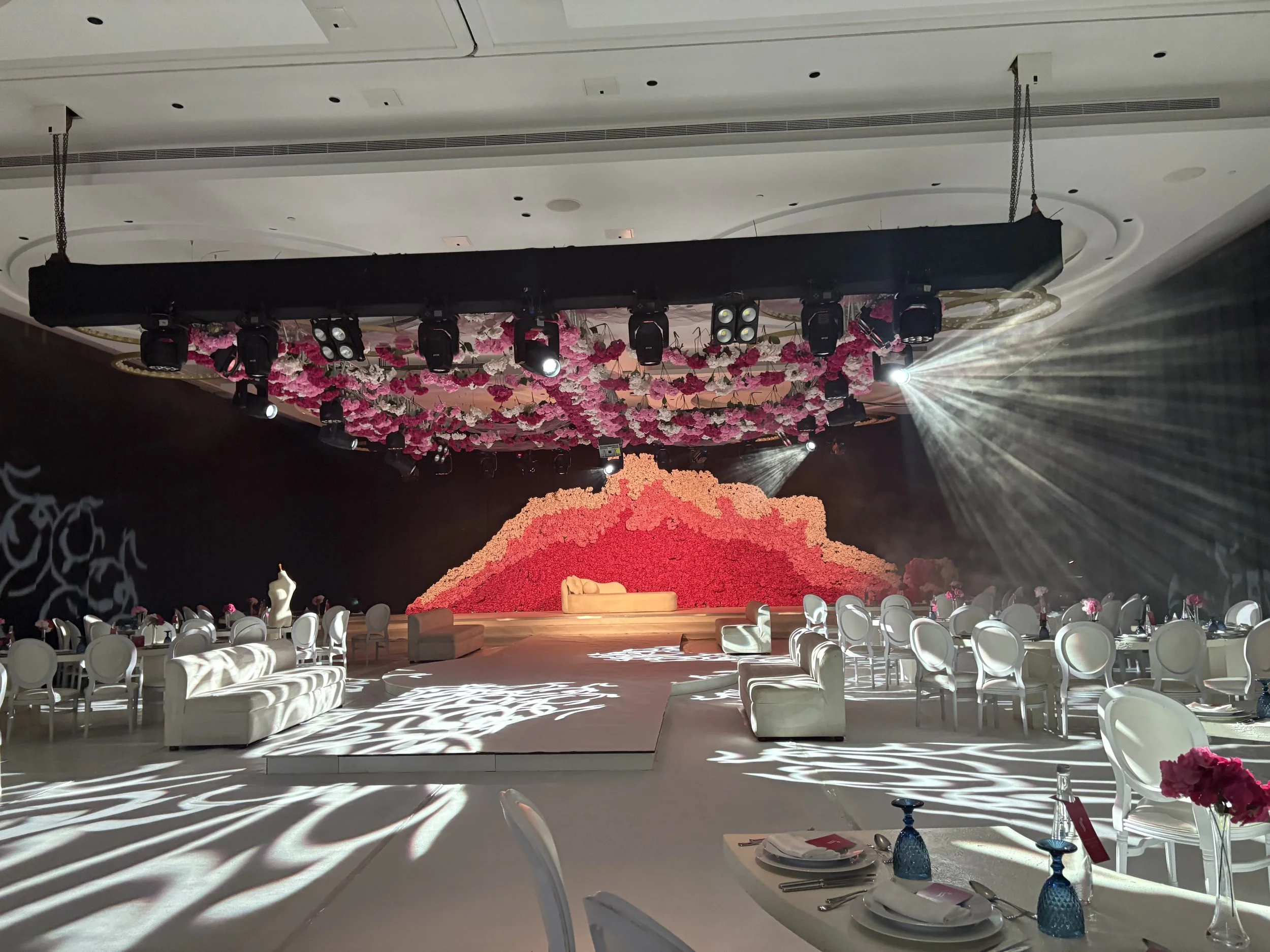 A decorated event stage with a floral backdrop, surrounded by round banquet tables and chairs, set for a celebration or wedding reception.