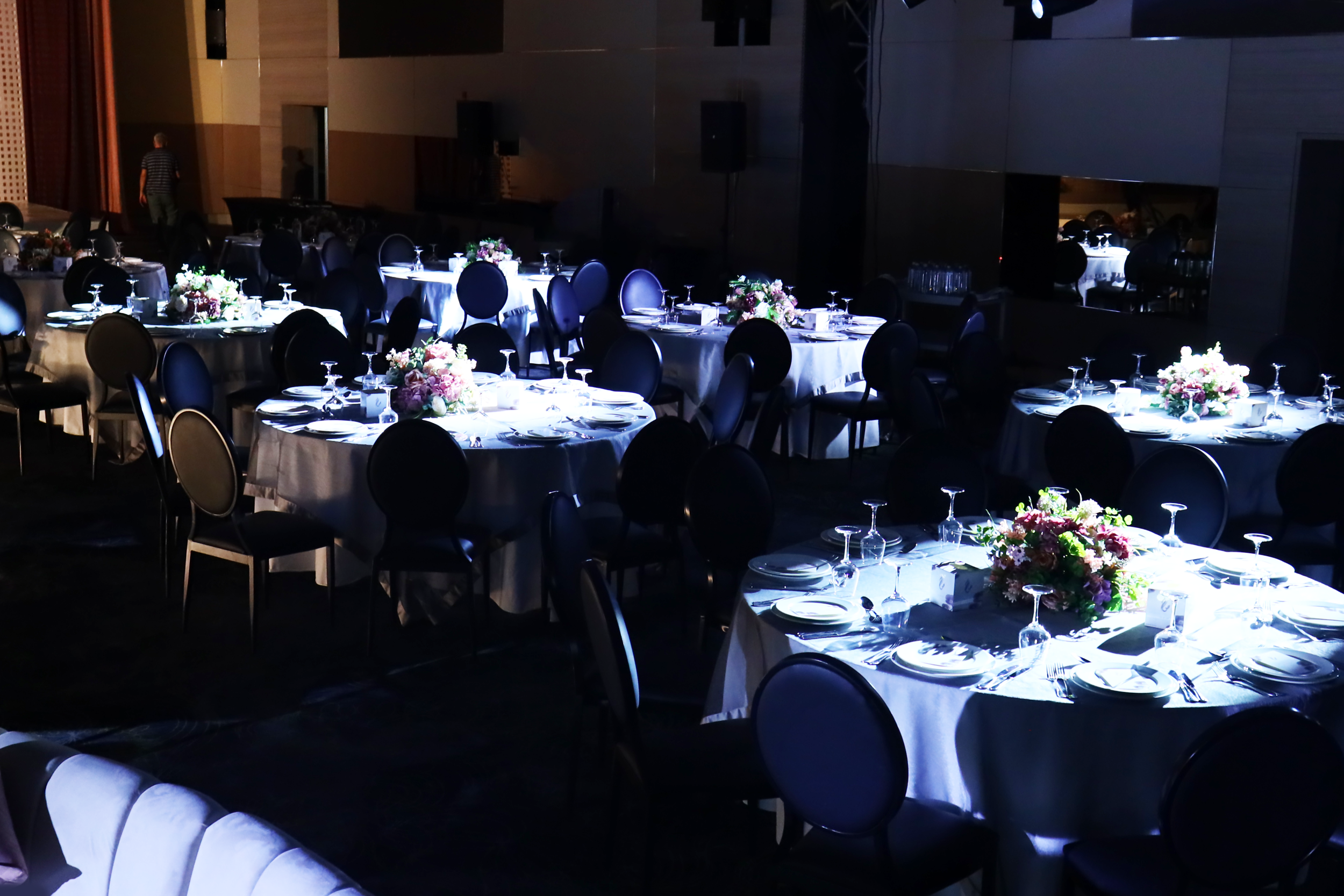 Dining hall with round tables covered in white tablecloths, decorated with floral centerpieces, set with plates, glasses, and silverware, illuminated with low lighting.