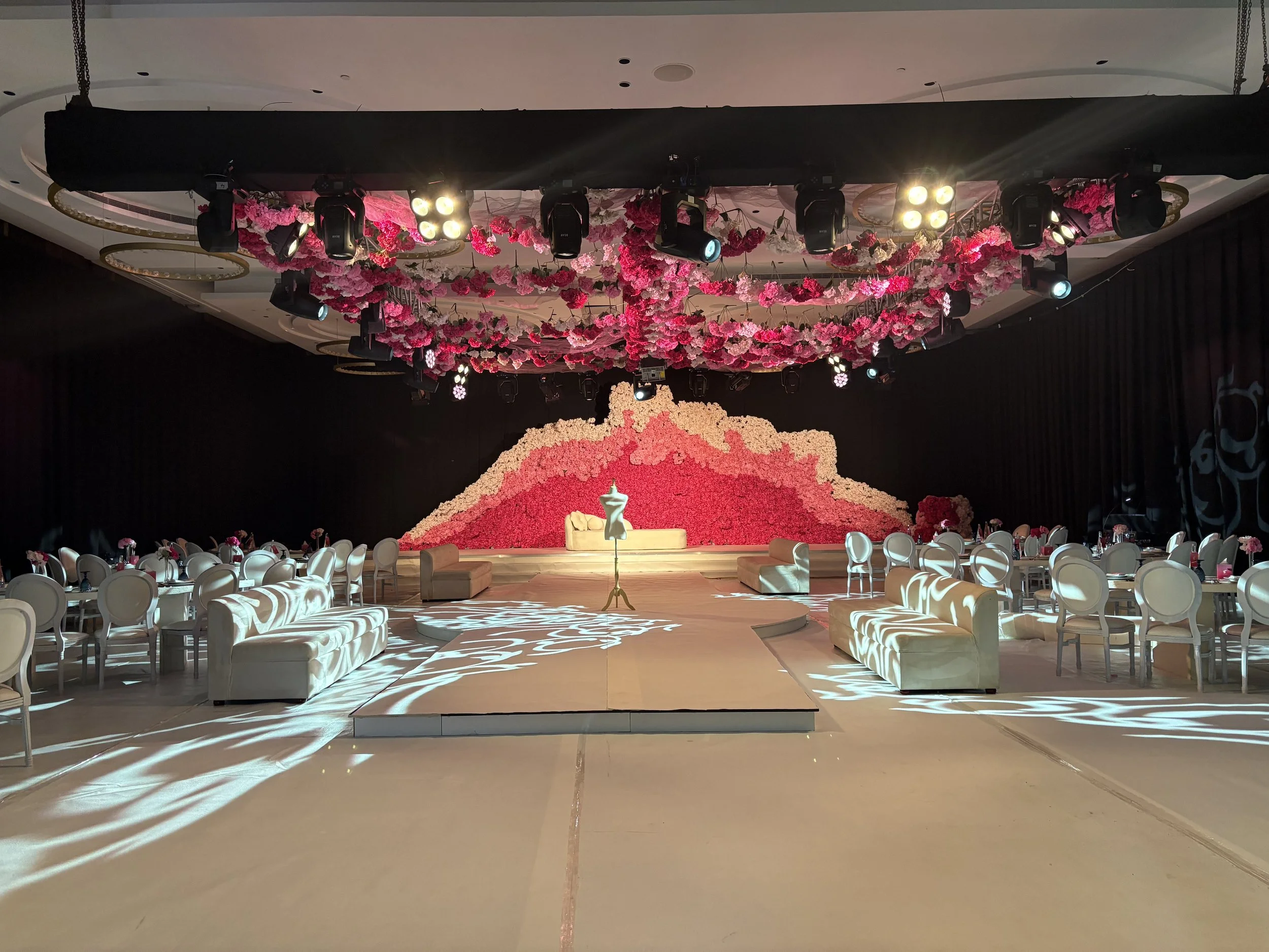 Event hall decorated with pink floral arrangements on the ceiling and a large pink and peach flower backdrop at the stage. White chairs and couches are arranged around the stage, and decorative lighting creates shadows on the floor.