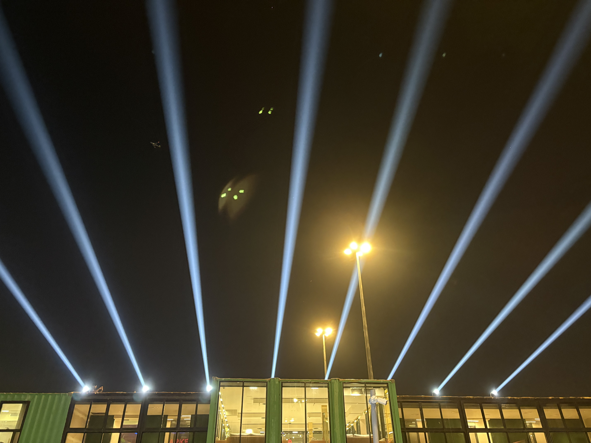 Night scene of an outdoor area with bright white and blue light beams shooting into the dark sky, illuminating the area. There are tall streetlights casting yellow light and a structure with large windows in the foreground.