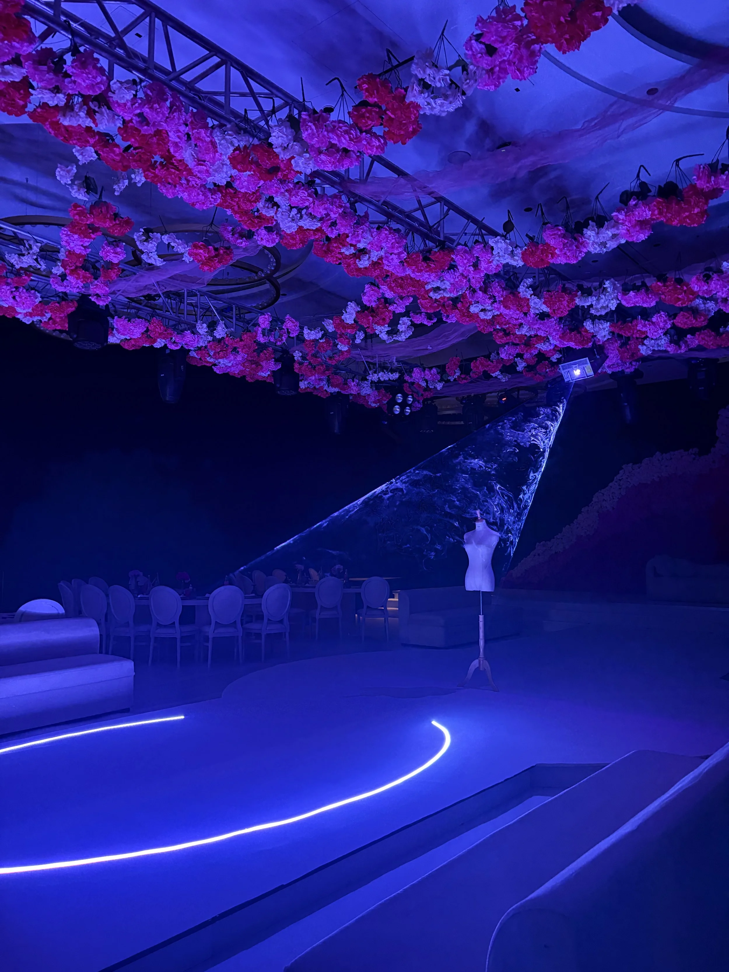 A dimly lit event space decorated with pink and white flowers hanging from the ceiling. A spotlight illuminates a mannequin on a stand and a long table with chairs in the background.