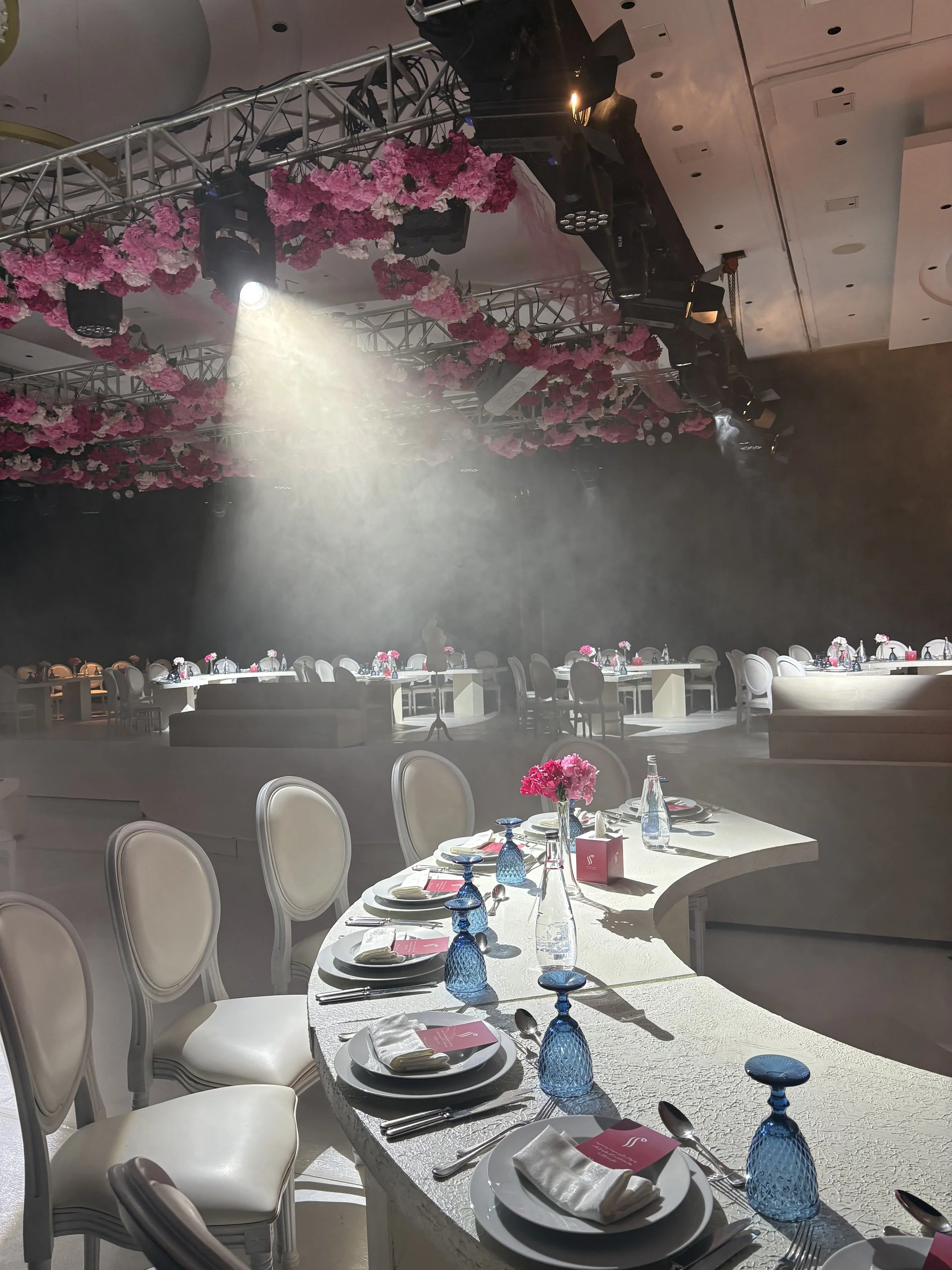 Event hall decorated with pink flowers and set for dining, with tables, chairs, and lighting fixtures suspended from the ceiling.