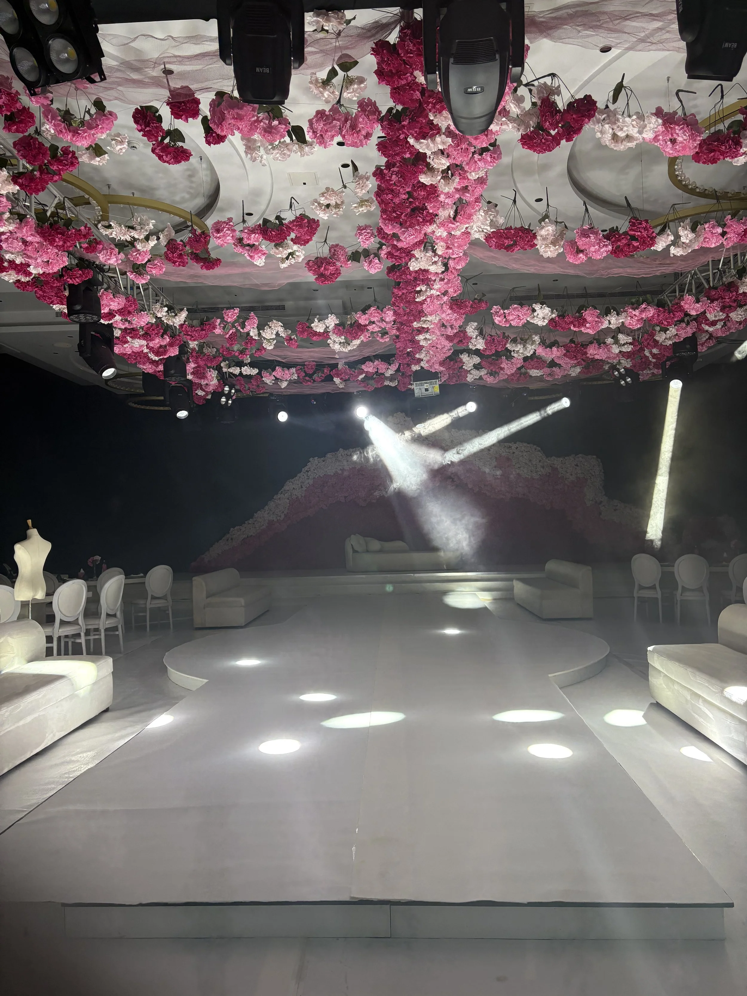 Interior of a decorated event hall with a white stage, pink and white floral hanging decorations on the ceiling, and white furniture including sofas and chairs, with stage lighting effects.