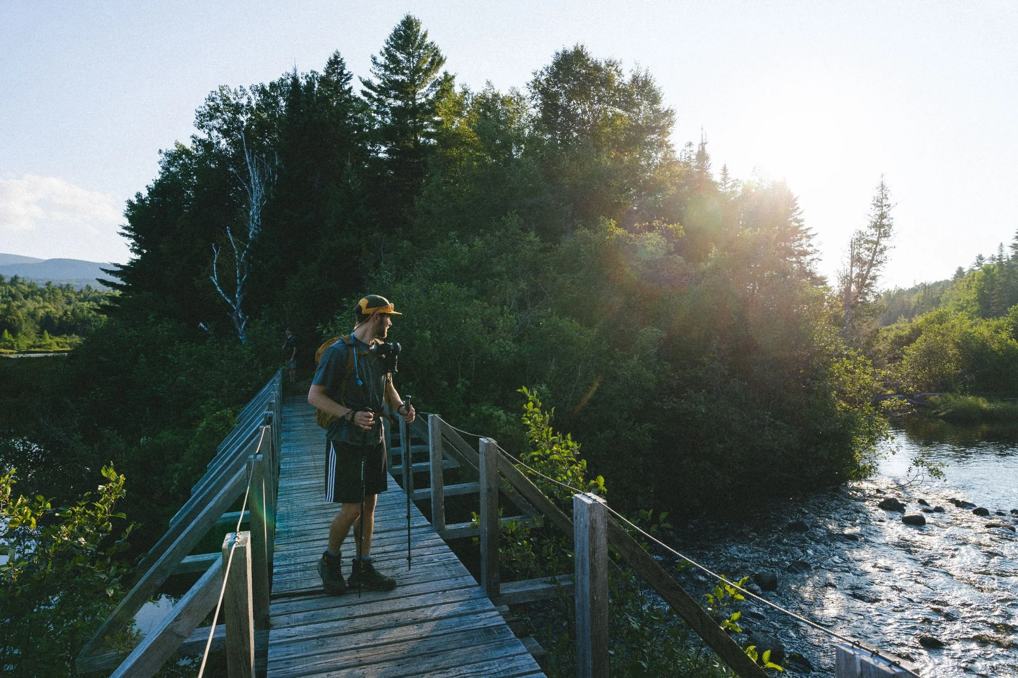 A person stands on a wooden bridge over a river, surrounded by lush green trees, with sunlight shining from the right side.