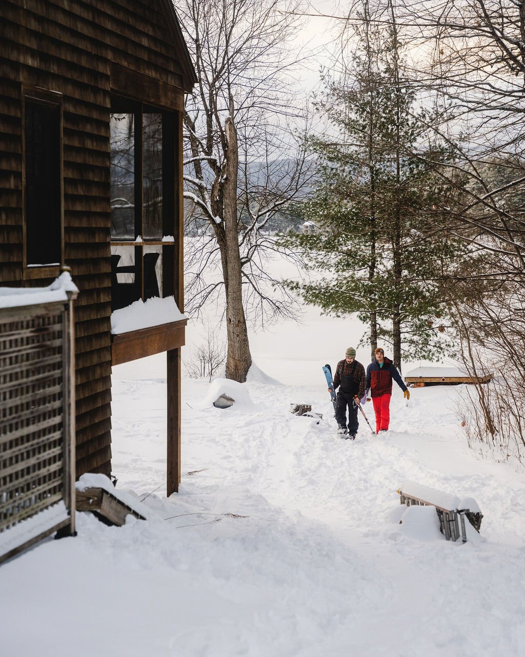 Two people walking through a snowy landscape near a wooden house, with trees and a snow-covered ground.