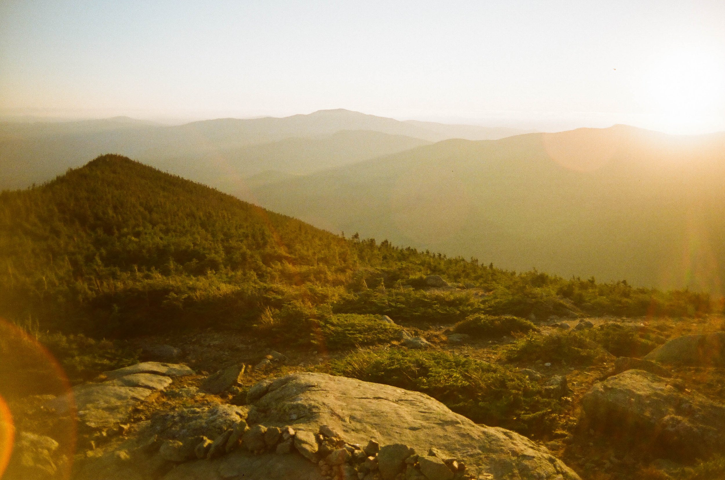 A mountainous landscape during sunset with layers of hills and a misty sky, rocks and vegetation in the foreground.