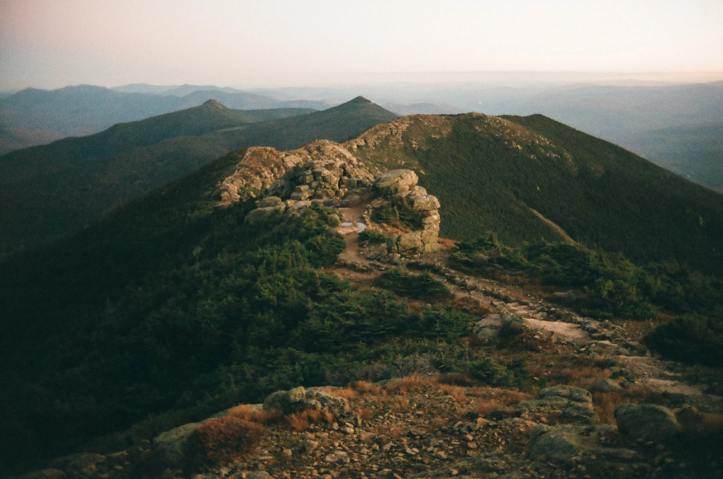 Mountain landscape with rocky peak and green vegetation, distant mountain range, and soft lighting at dawn or dusk.