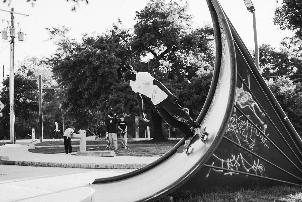A skateboarder riding on a large curved ramp in a park, with three people in the background near trees and a sidewalk.