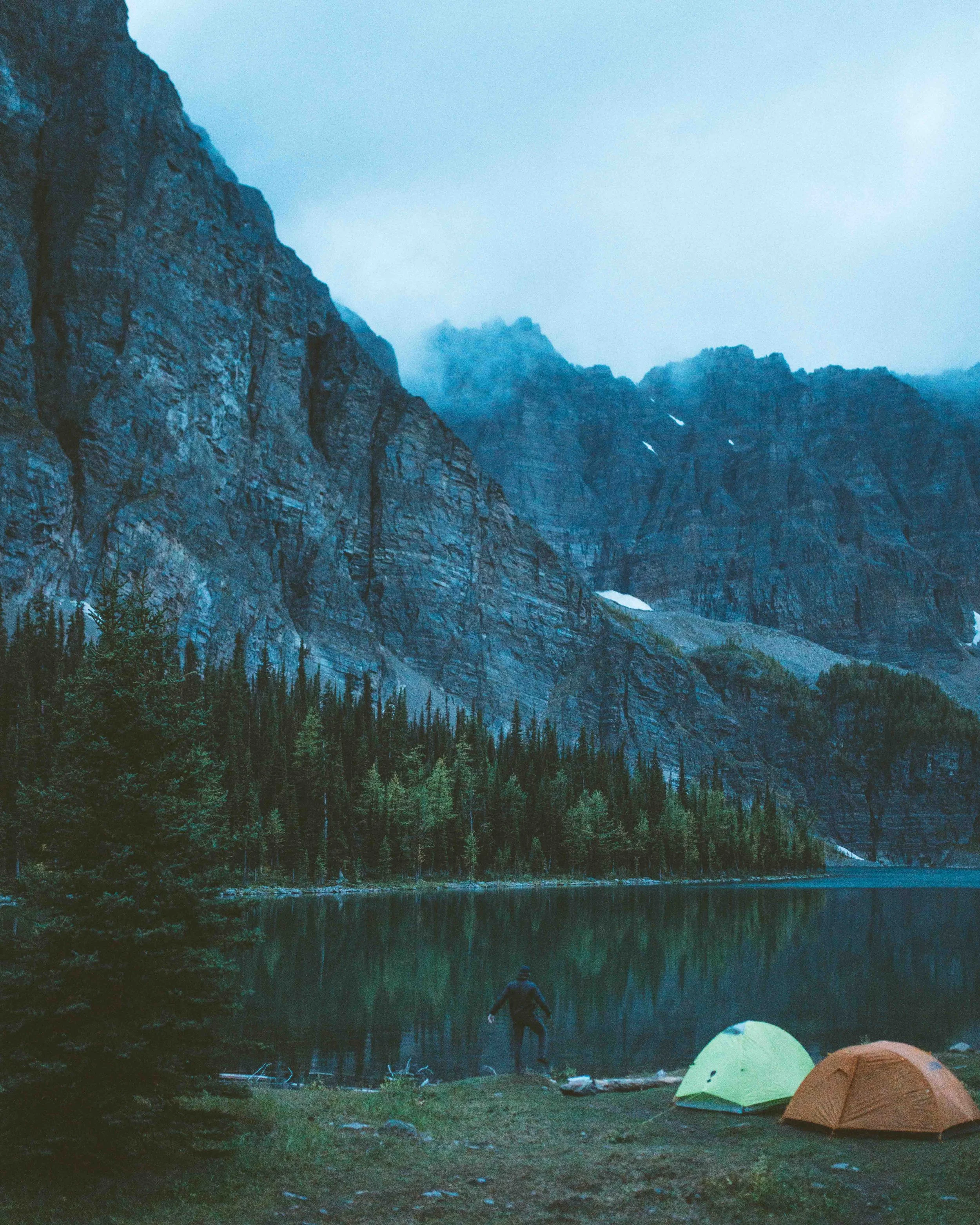 A person standing by a lake with mountains and trees in the background, and two tents pitched near the water's edge.