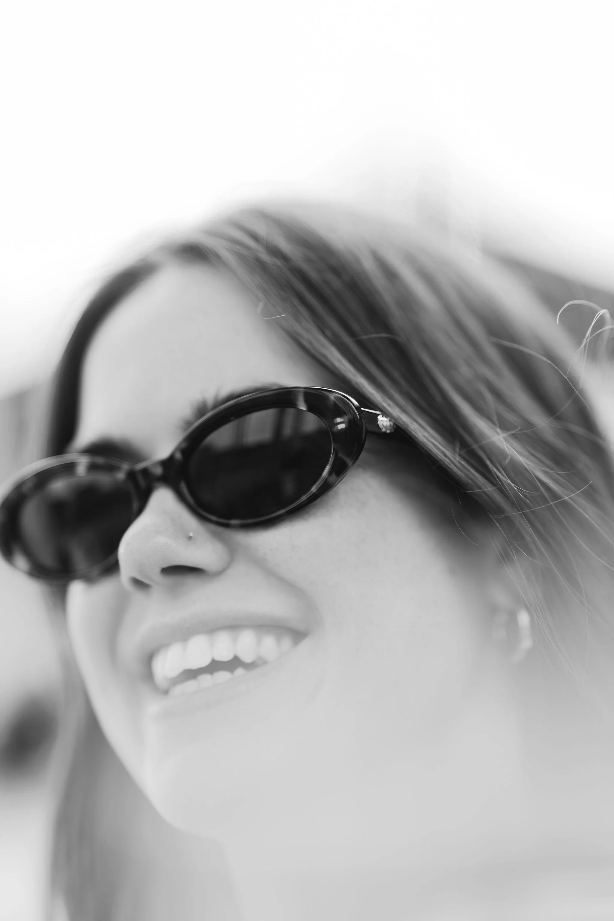 Close-up black and white photo of a woman with sunglasses and a nose piercing, smiling.