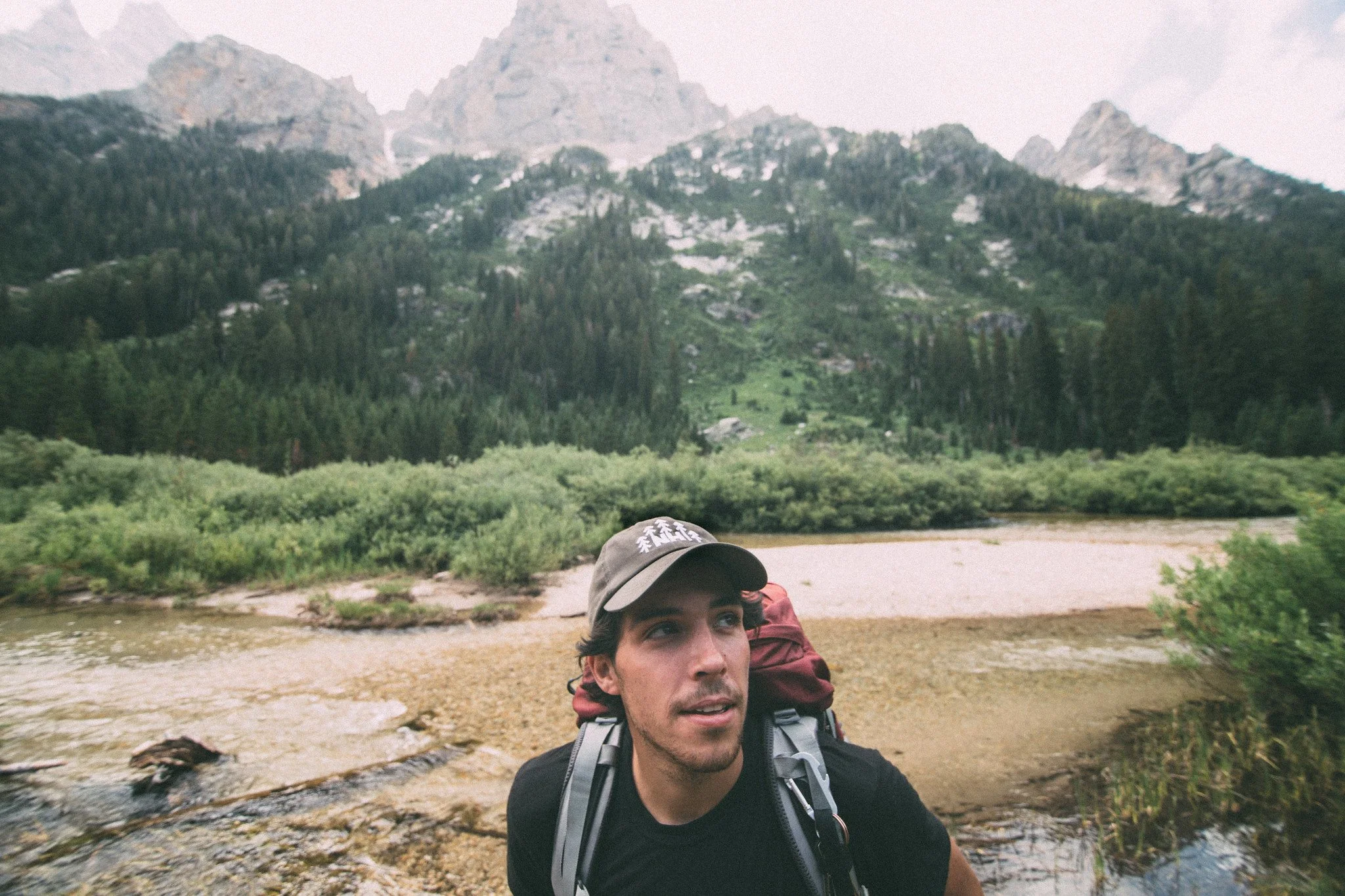 A young man with a backpack hiking outdoors, with mountains and a river in the background