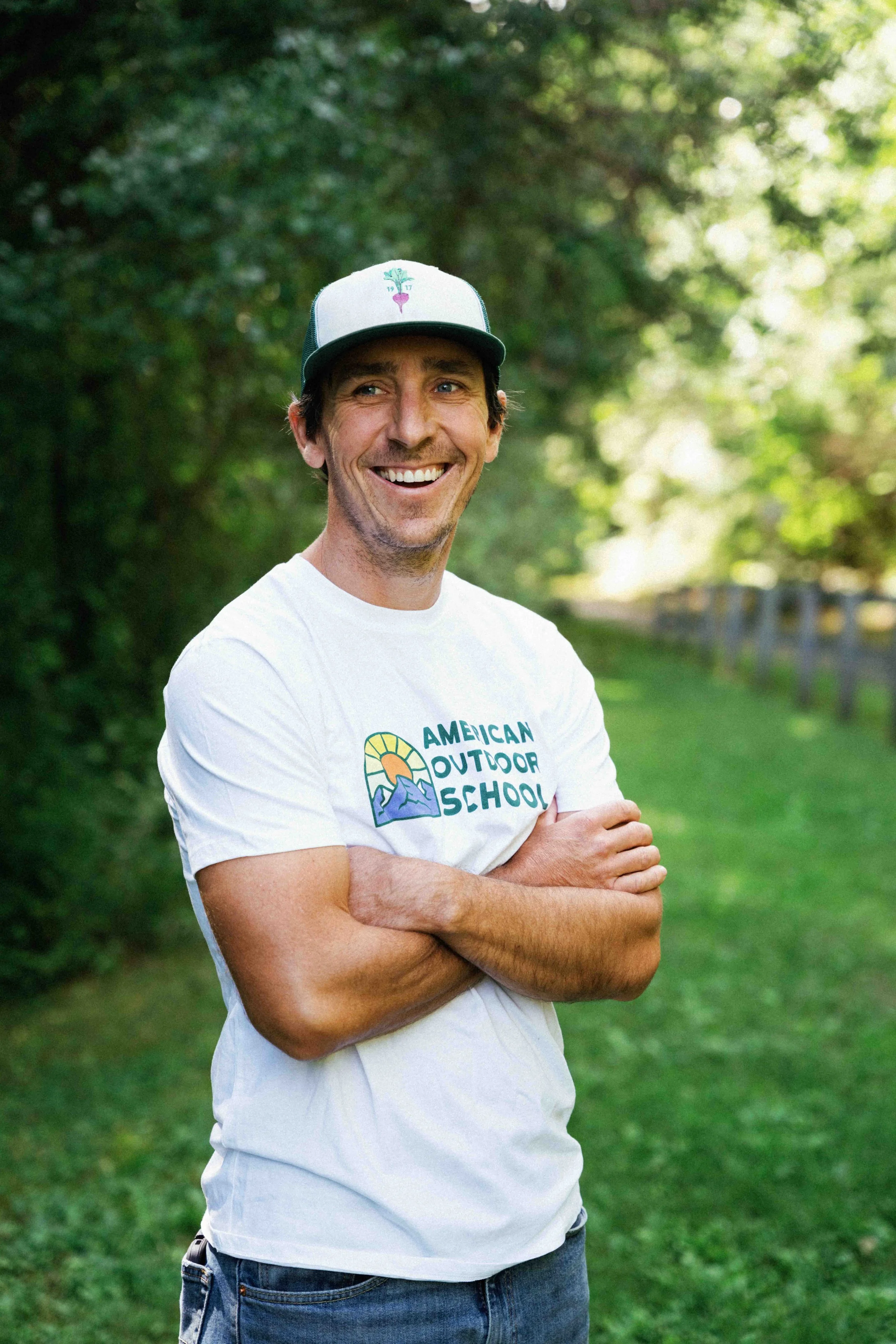 A man standing outdoors in a park-like setting with trees and grass, smiling, with arms crossed, wearing a white t-shirt with 'American Outdoor School' logo and a trucker cap.