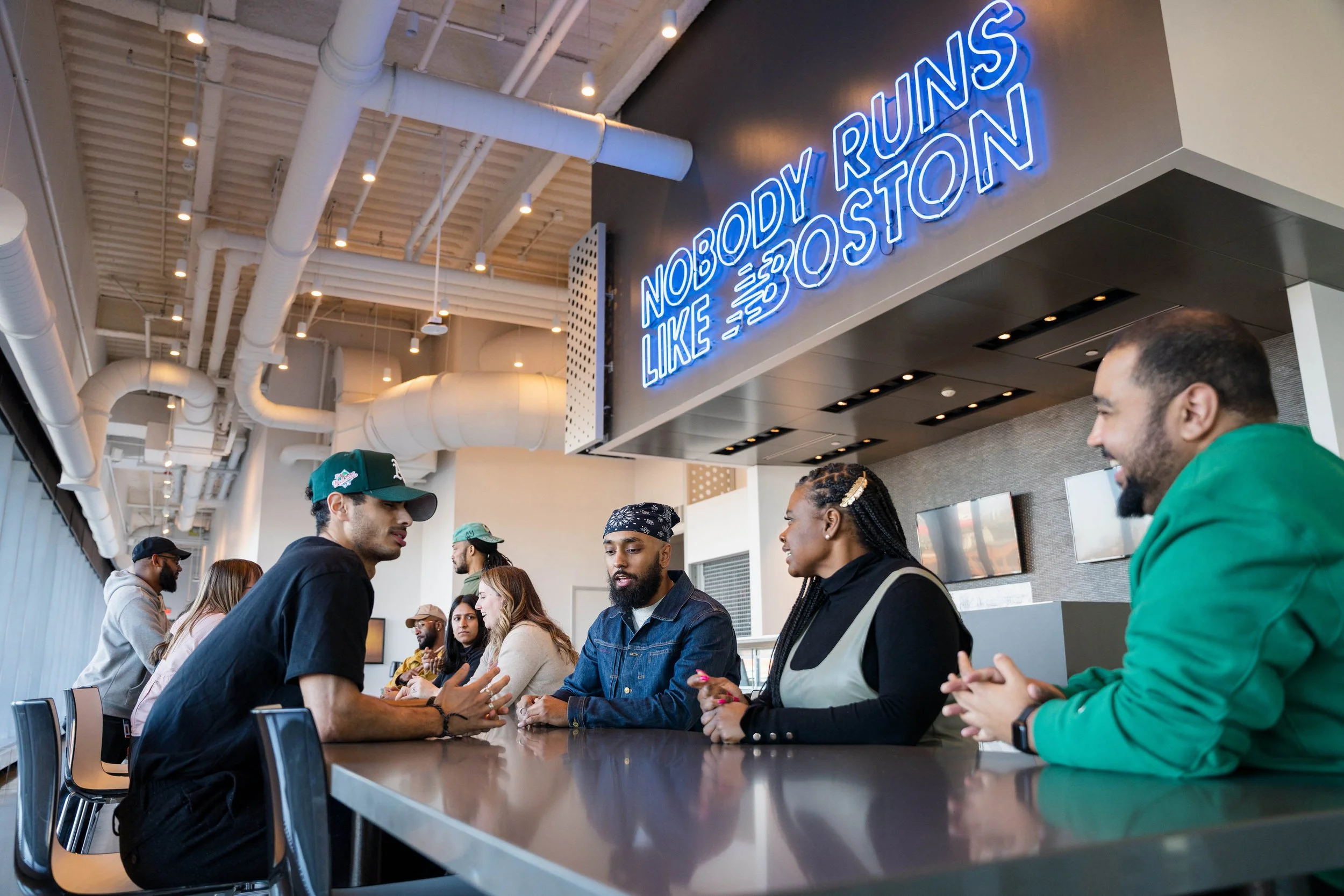 Group of diverse people sitting at a counter, talking, with a neon sign that says 'Nobody Runs Like Boston' in the background.