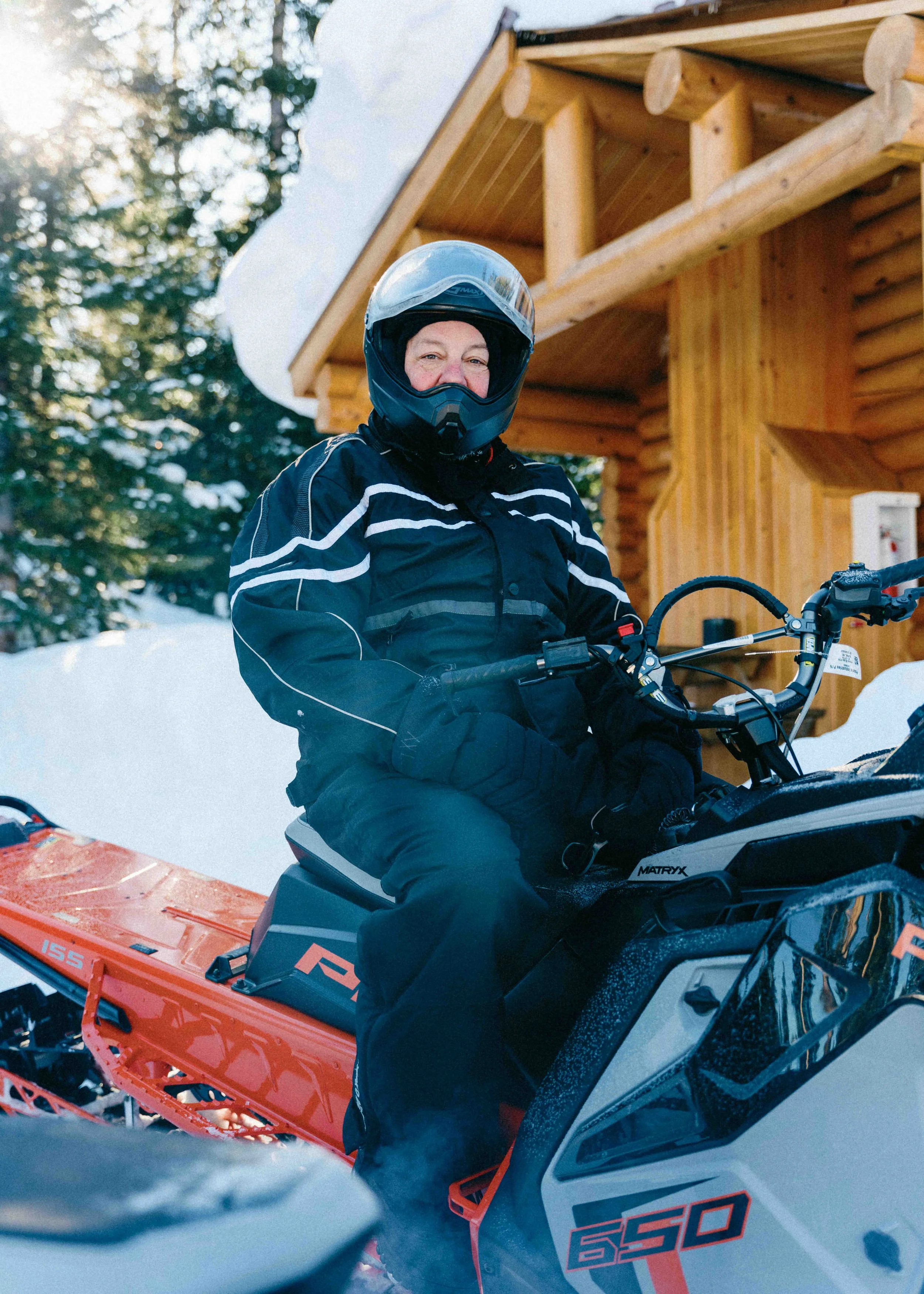 A person in ski gear sitting on a snowmobile outdoors in a snowy, wooded area with a wooden cabin in the background.