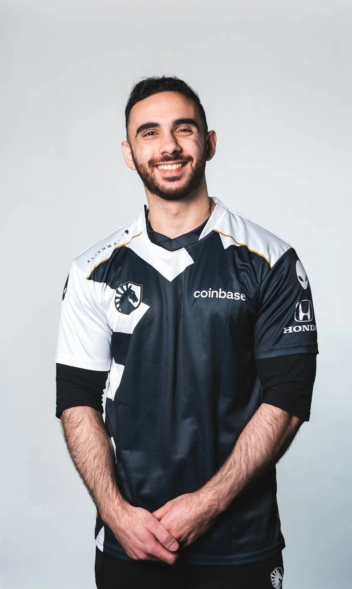 A young man with dark hair and a beard smiling, wearing a black and white esports jersey with team and sponsor logos, standing against a plain light gray background.
