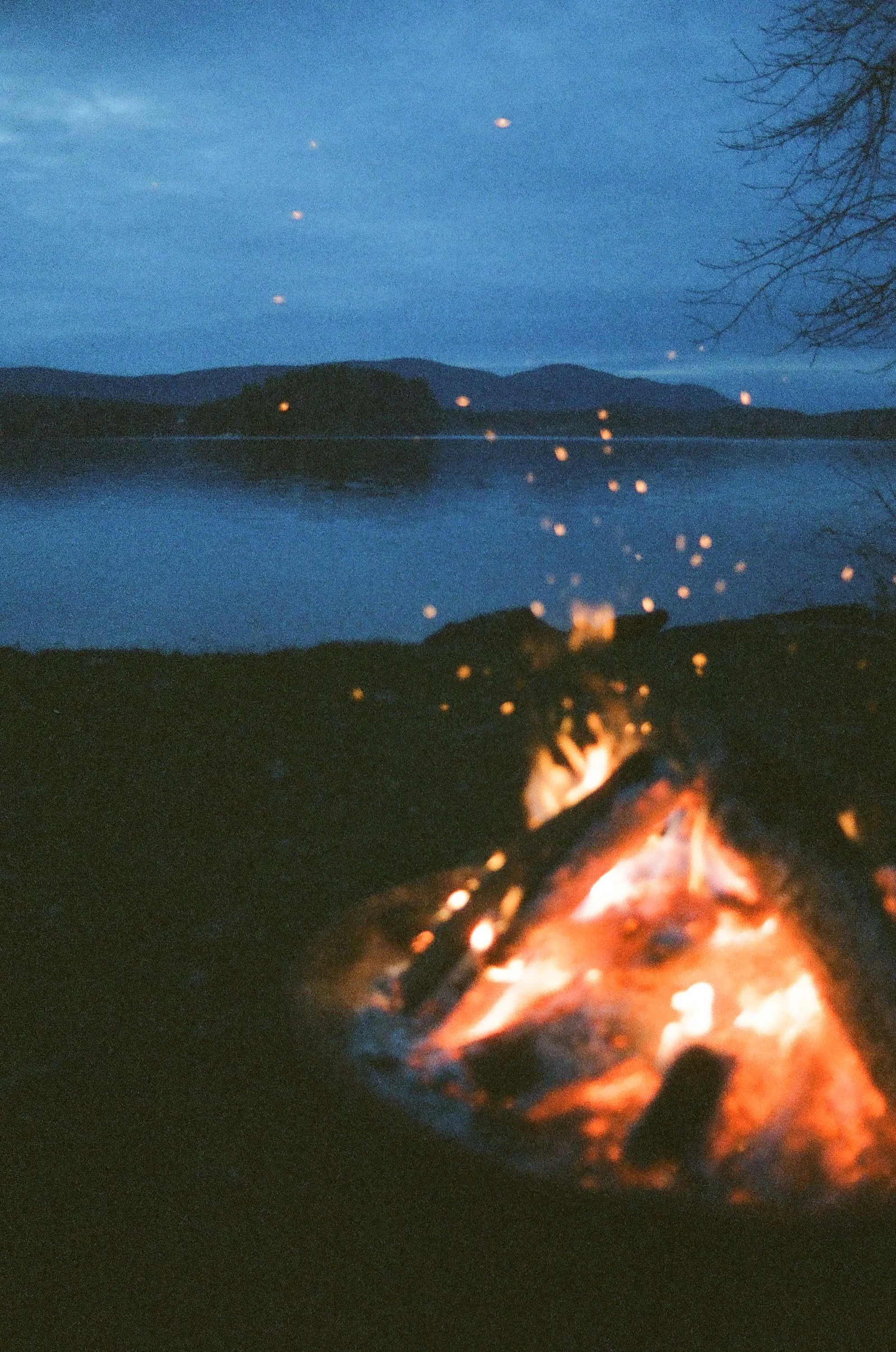 A campfire near a body of water with mountains in the background, taken during dusk, with floating sparks in the air.