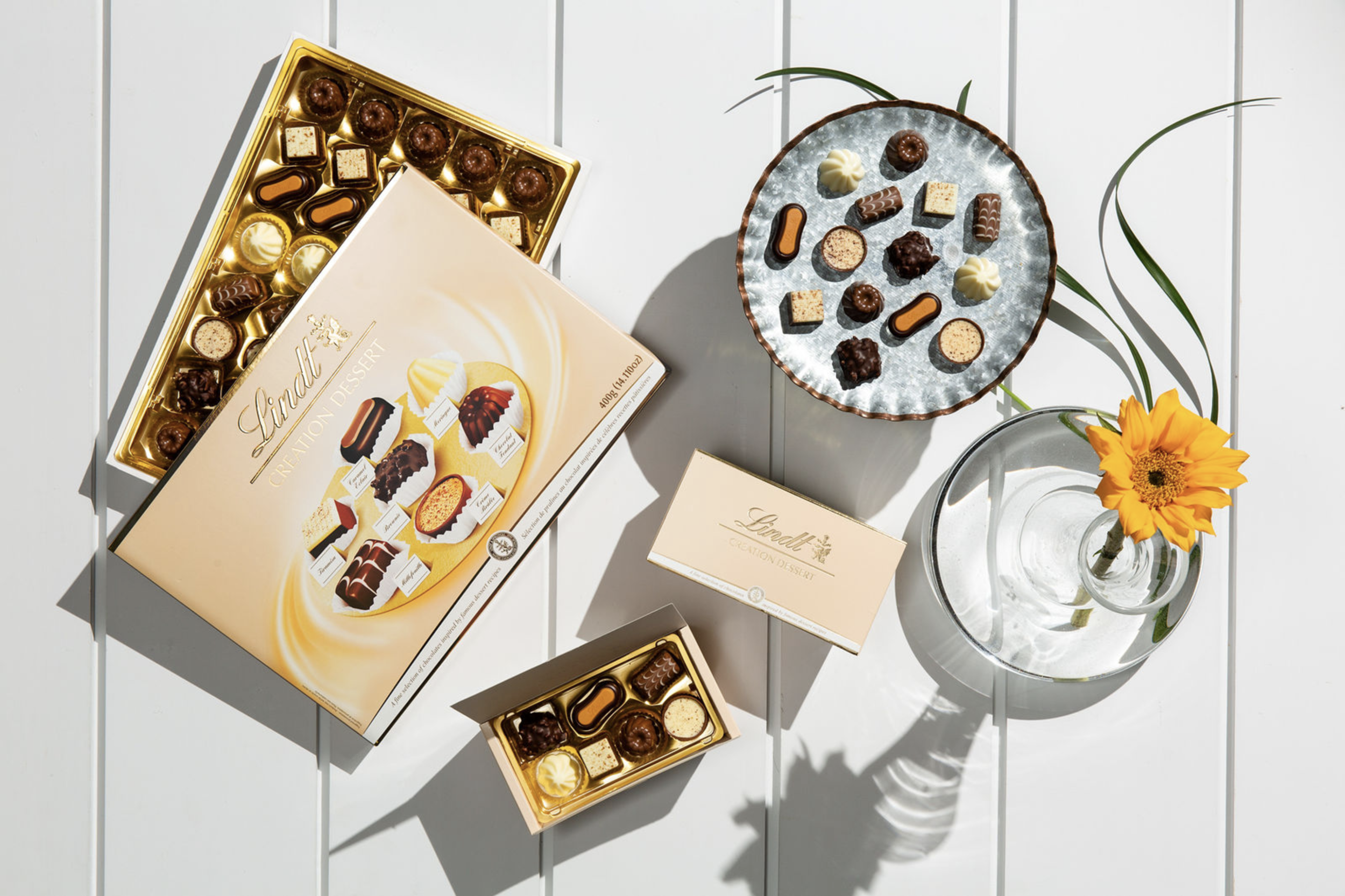 Lindt creation dessert box with assorted chocolates, a small tin of chocolates, a round plate with chocolates, and a glass vase with a yellow flower all arranged on a white wooden table.