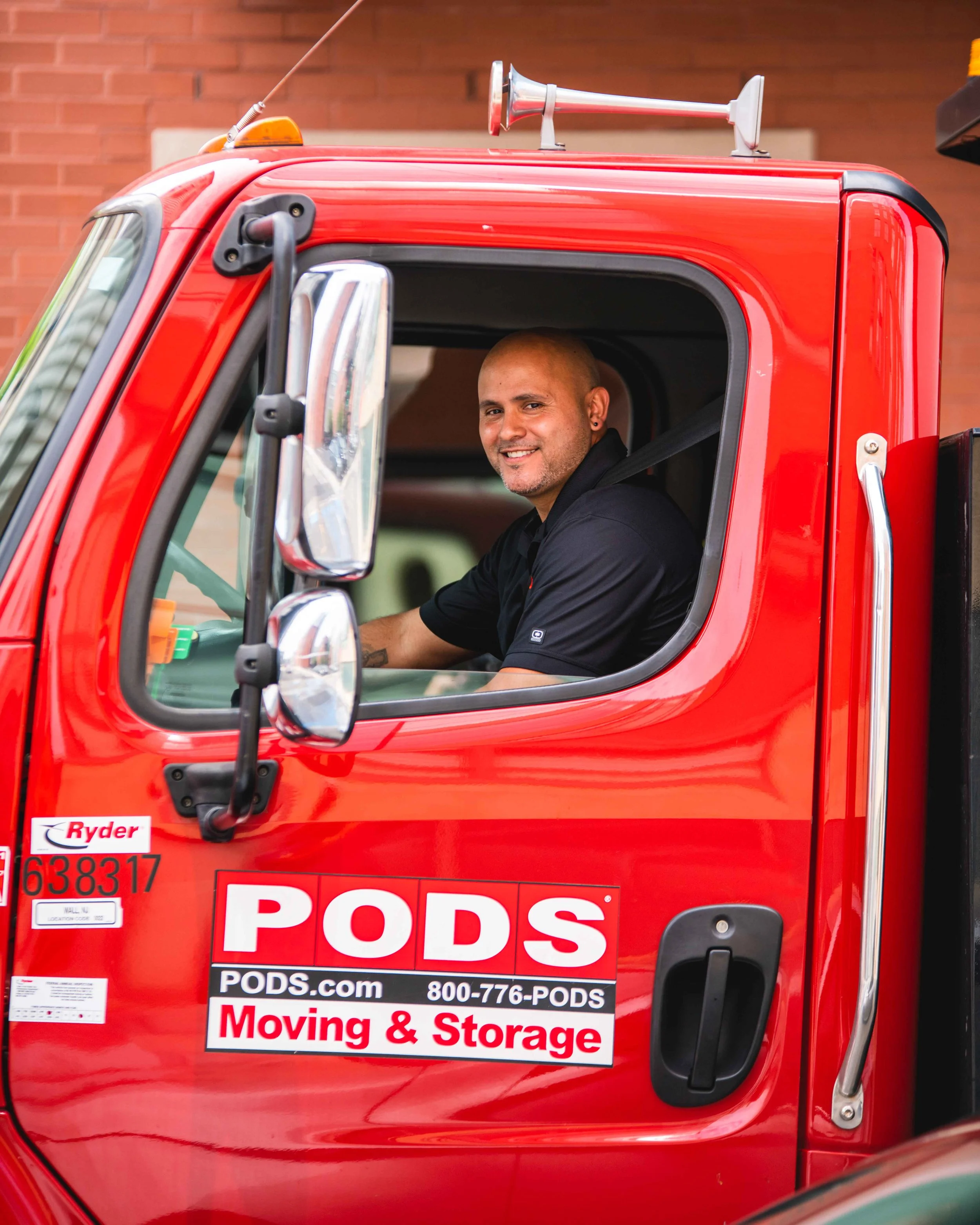 A smiling man is sitting in the driver’s seat of a red PODS moving truck, looking out the window. The truck is parked outside a building with a brick wall.