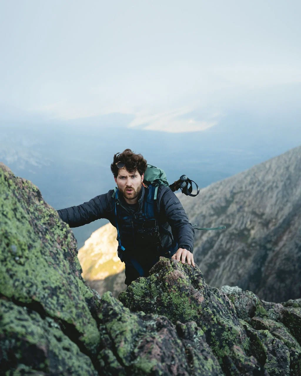 A man climbing rocky, moss-covered mountain terrain with a panoramic mountain landscape in the background.
