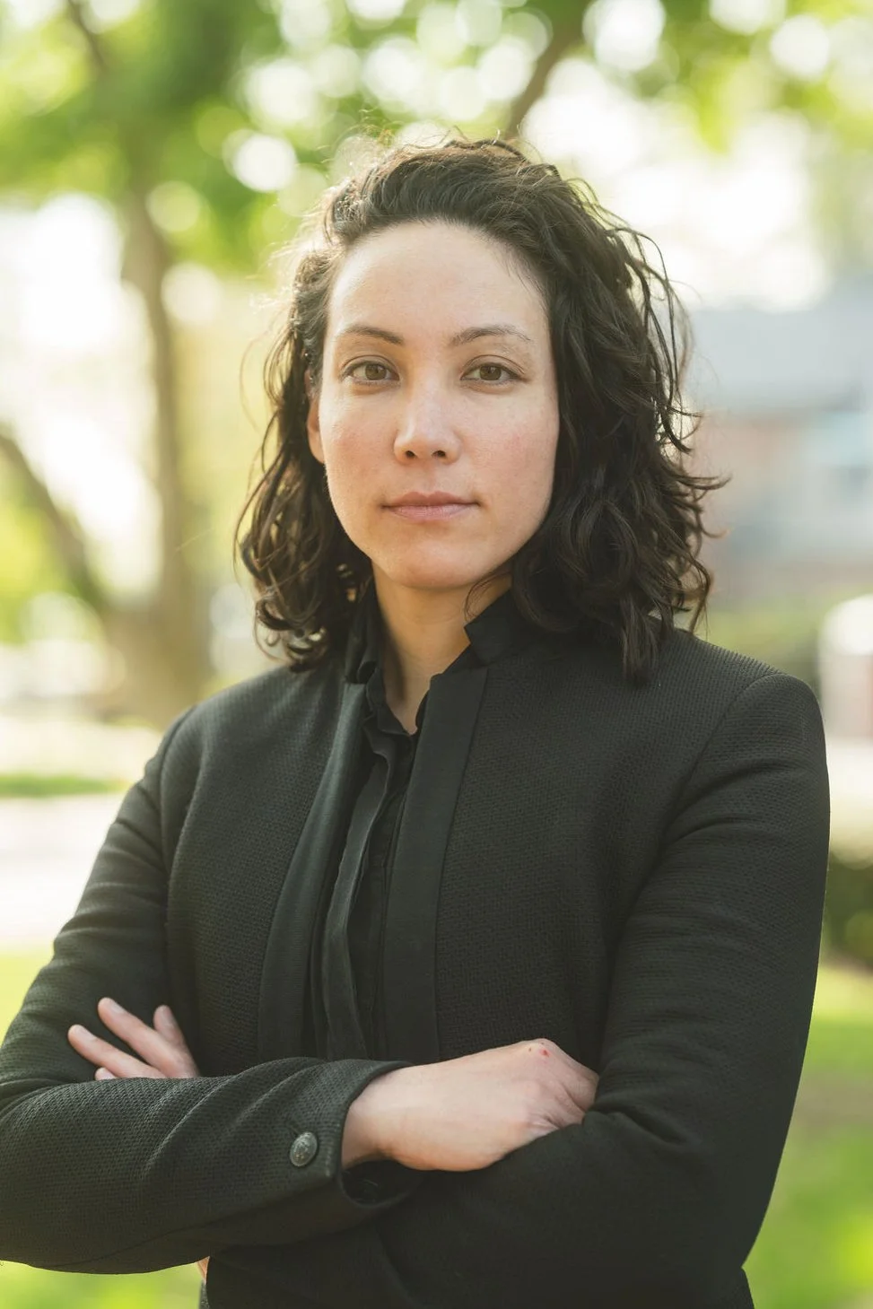 A woman with dark, curly hair standing outdoors with her arms crossed, wearing a black blazer and blouse, with a blurred background of trees and sunlight.