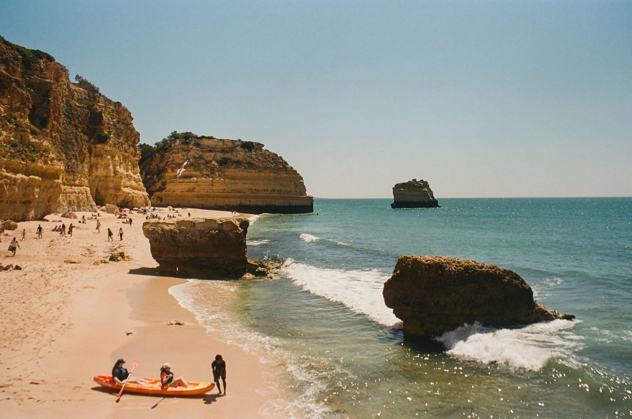 Beach with large rocks, cliffs, and a blue ocean. People are sunbathing and walking, with some preparing to kayak. A waterfall flows down one cliff side.