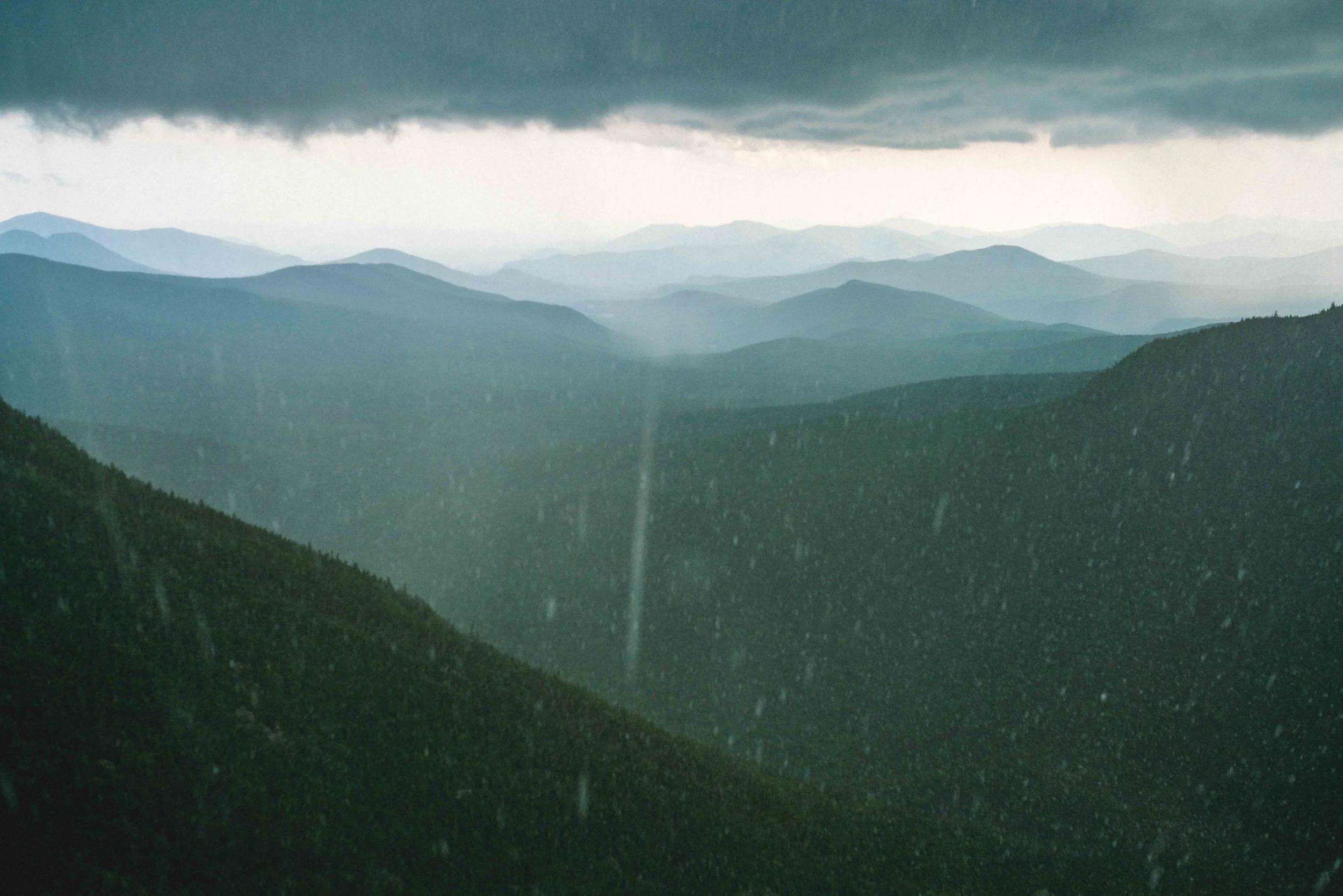 A mountain landscape with multiple layers of mountain ridges in shades of blue and green, under a dark, cloudy sky.