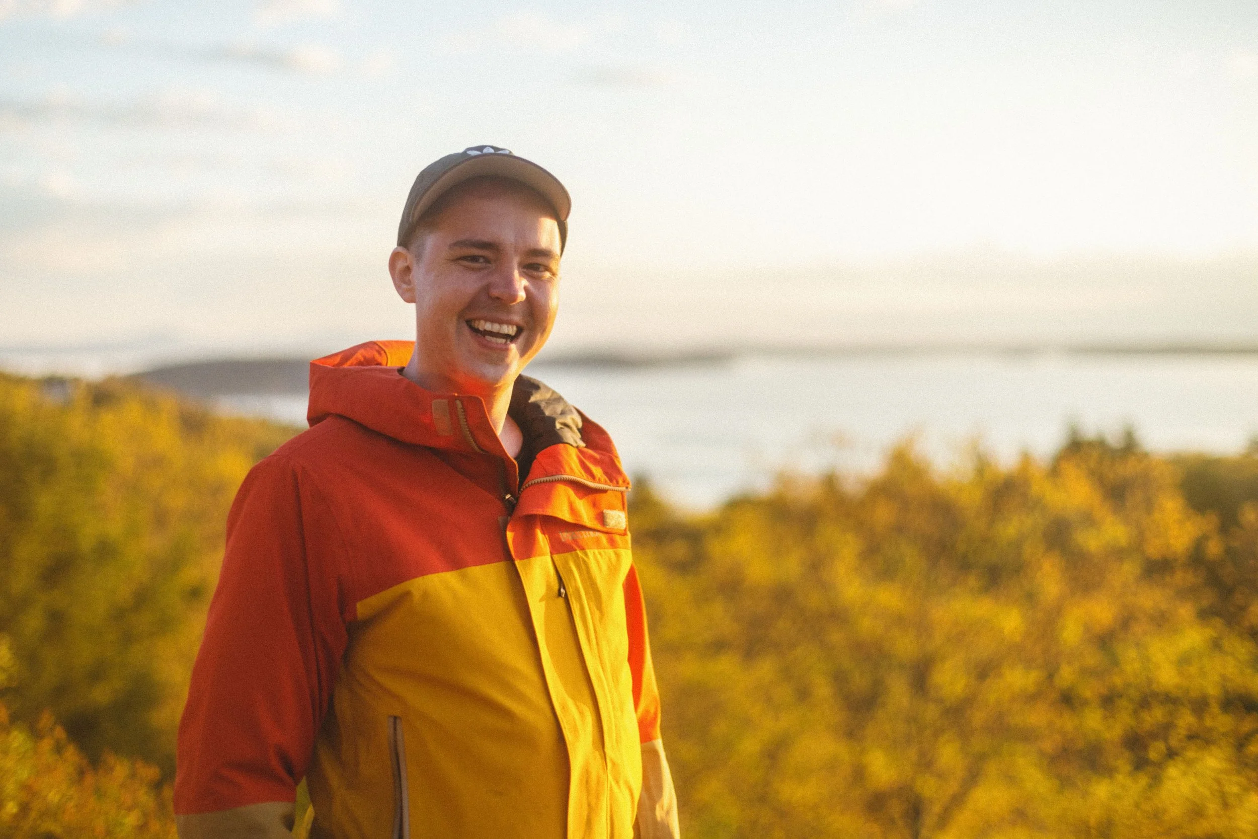 A young man outdoors smiling in a red and yellow jacket with trees and water in the background during sunset.