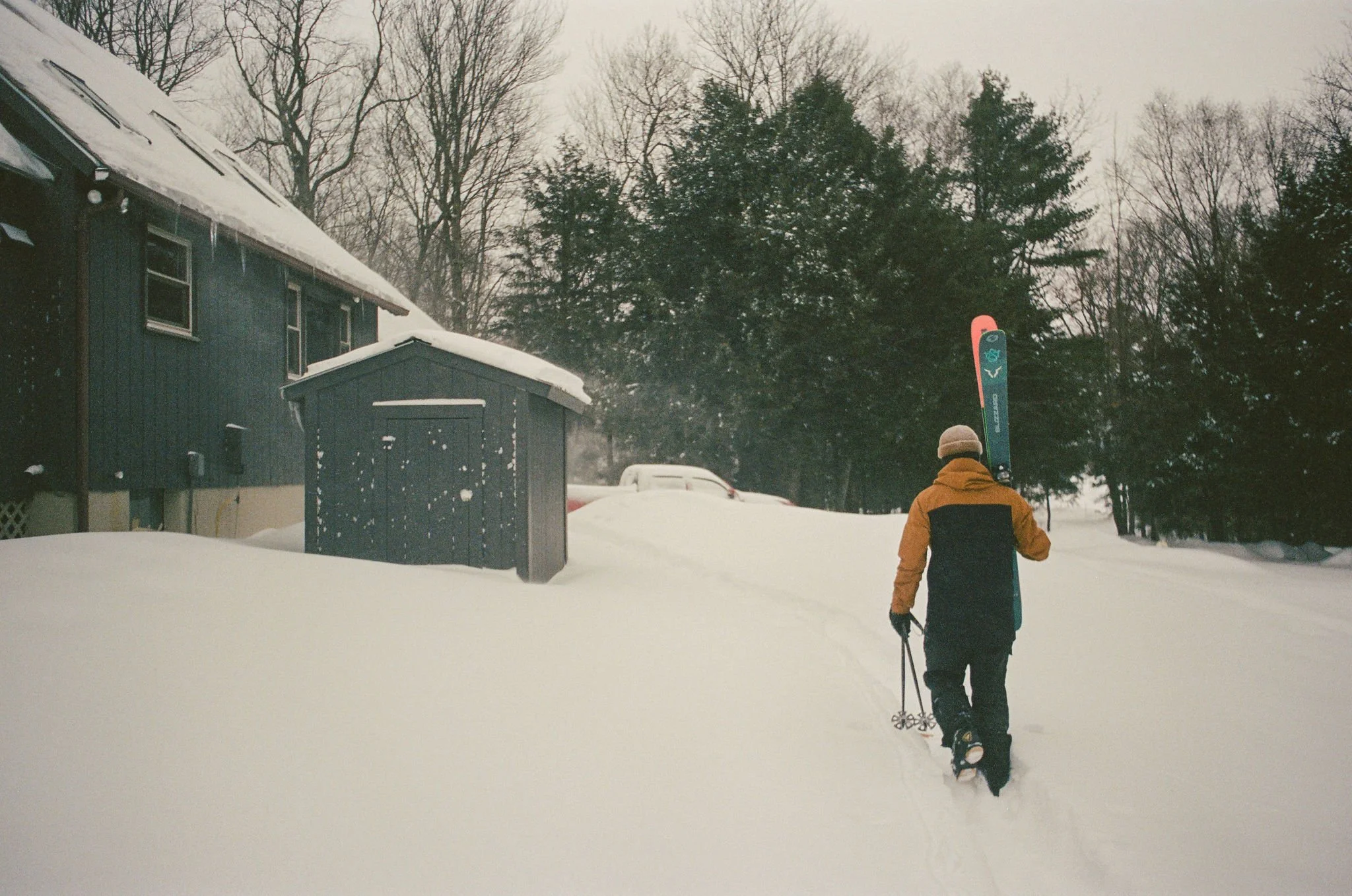 Person walking through deep snow while carrying a snowboard, dressed in winter clothing, near a blue house and shed in a snowy landscape with trees.