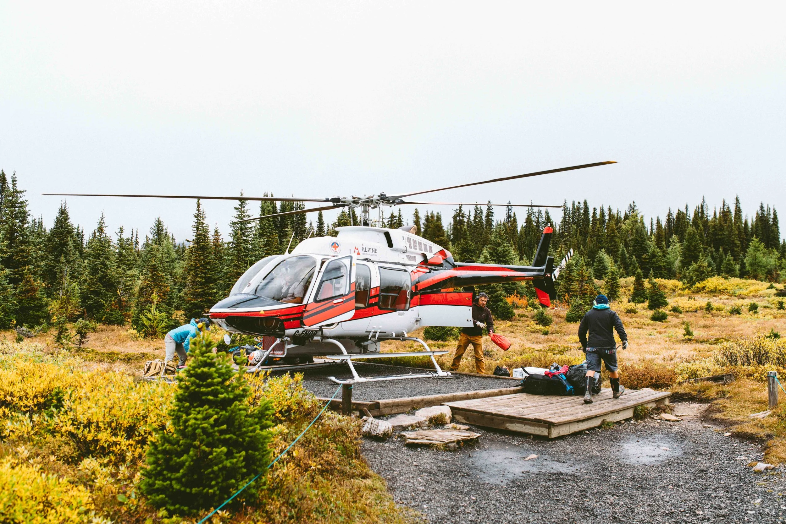 A rescue helicopter on a wooden landing pad in a forested area with three rescue workers preparing gear.