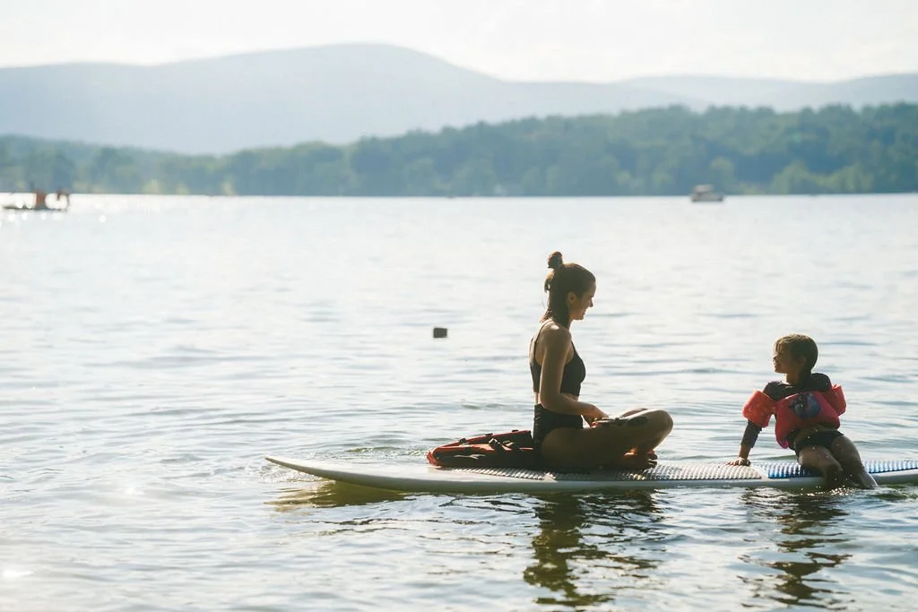 A woman and a young boy sitting on a paddleboard on a lake, with the woman facing the boy and the boy wearing orange flotation devices.