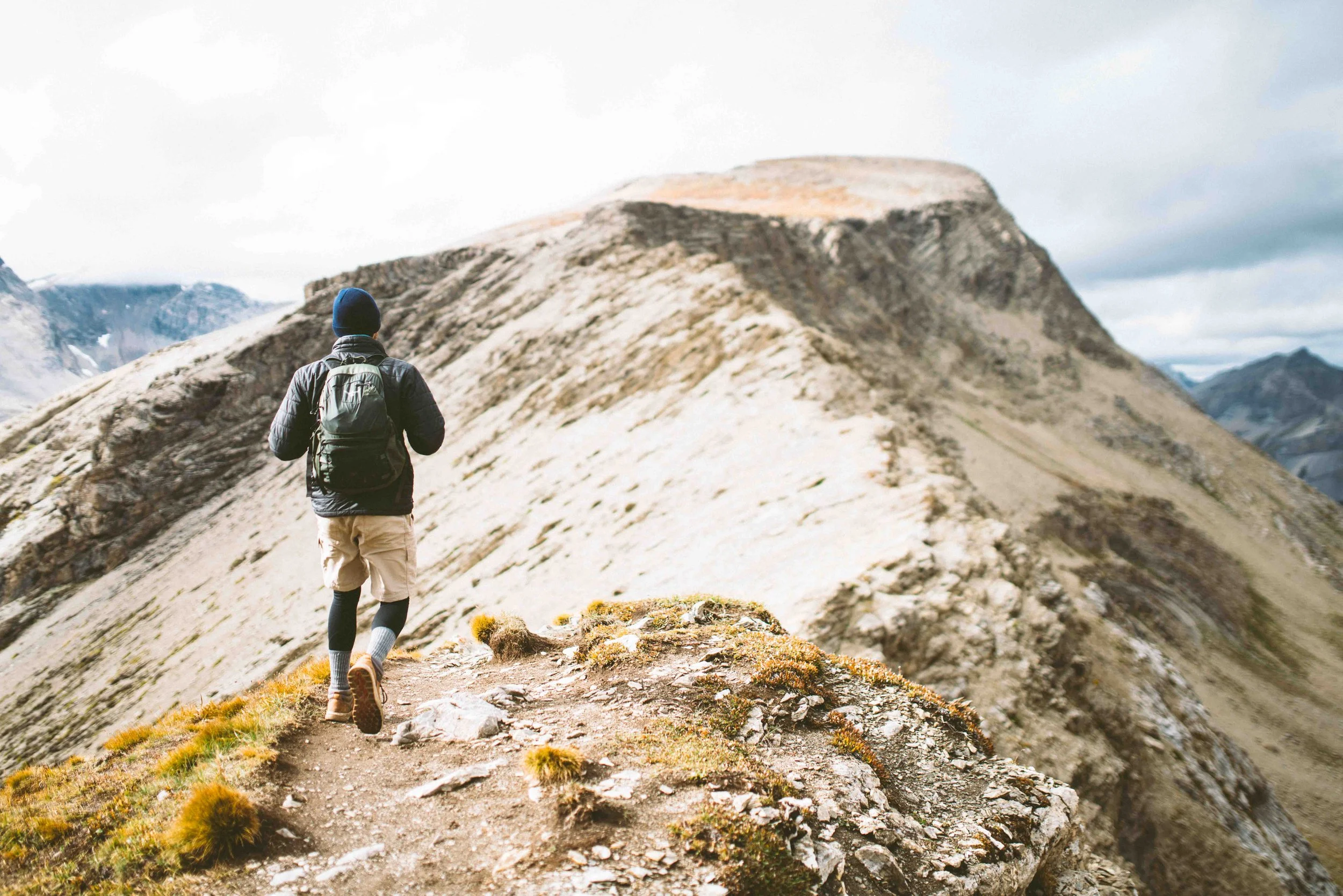 A hiker walks on a mountain trail surrounded by rocky terrain and patches of grass, with towering mountain peaks in the background under a cloudy sky.