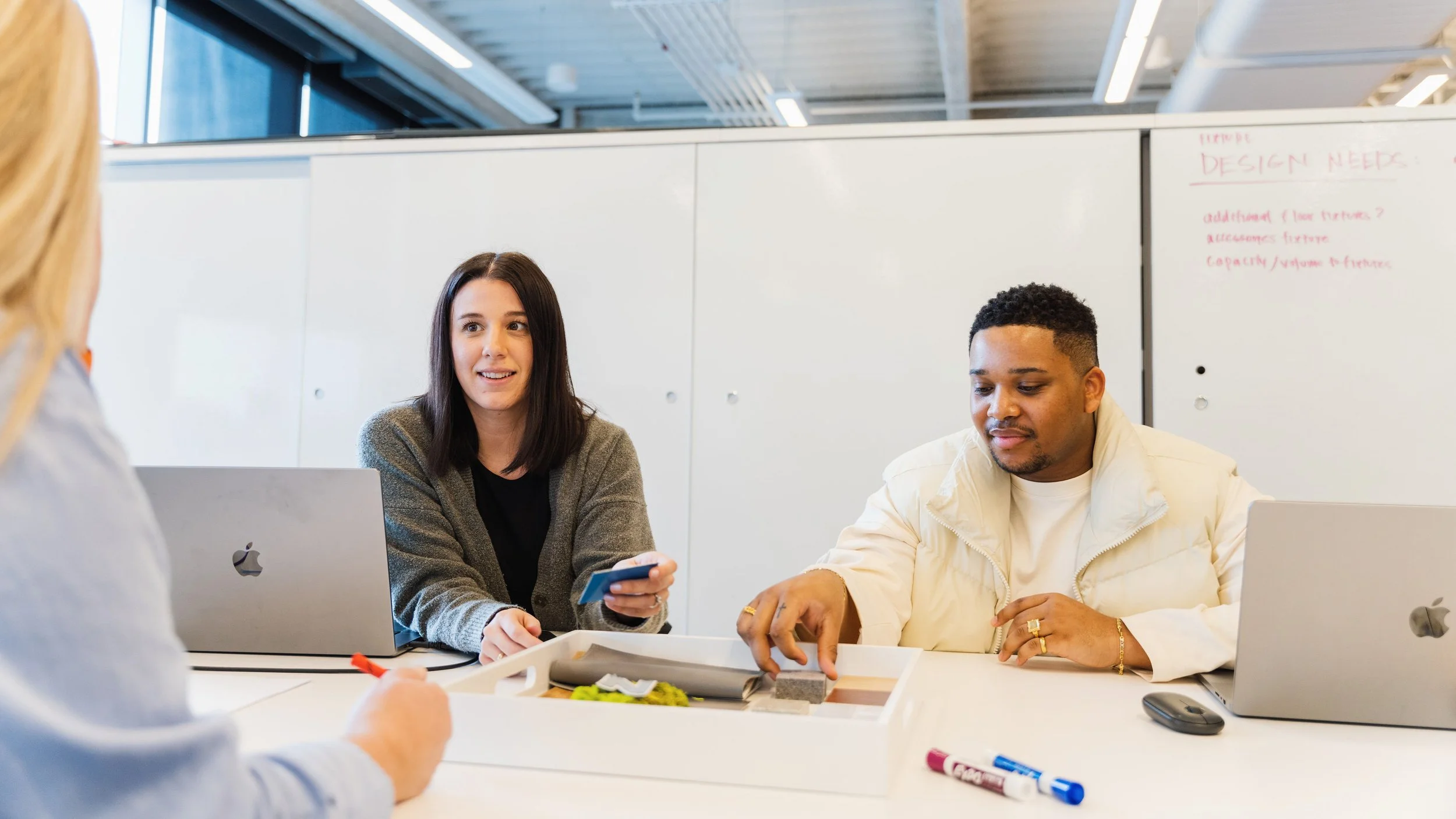 Three people engaged in a meeting at a conference table, with laptops and design samples, in a modern office setting.