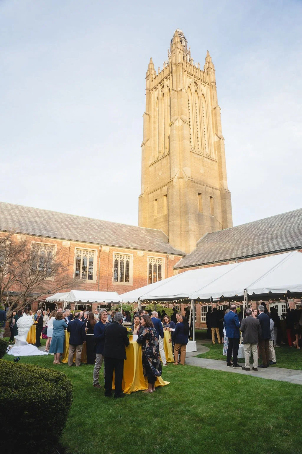 People gathered outdoors near a large historical brick building with a tall clock tower during a social event, with tents and greenery around.