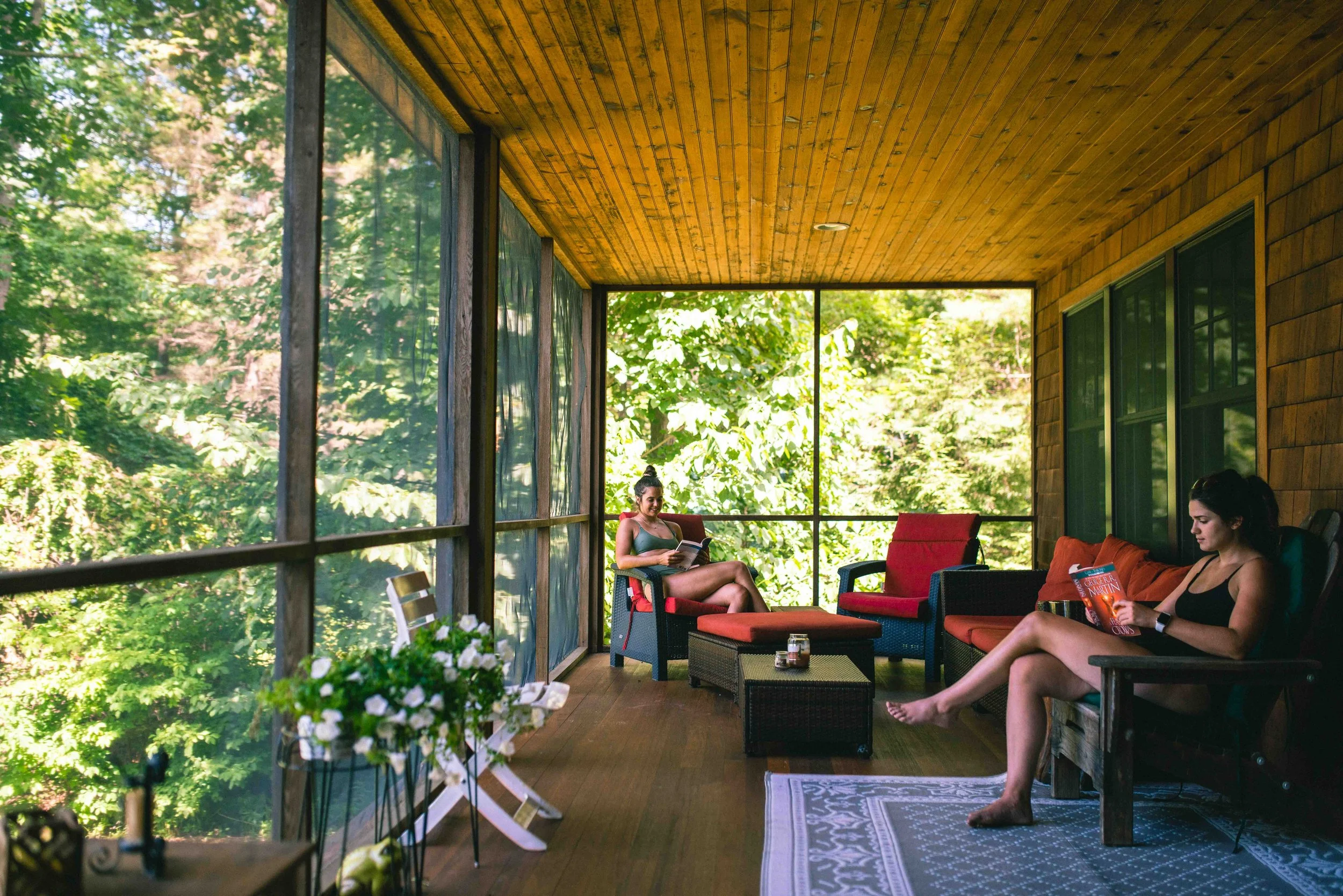 Two women relaxing on a screened porch with wooden walls and ceiling, surrounded by green trees, one reading a book and the other reading a magazine.