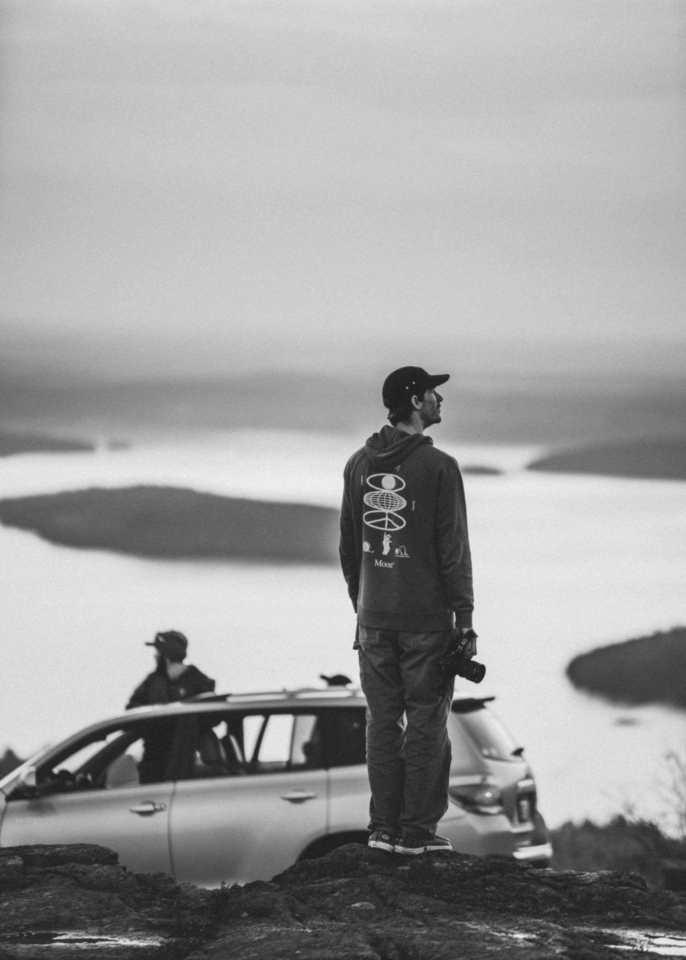 A man standing outdoors on rocks, holding a camera, with a car and another person in the background, overlooking a landscape with water and islands, in black and white.