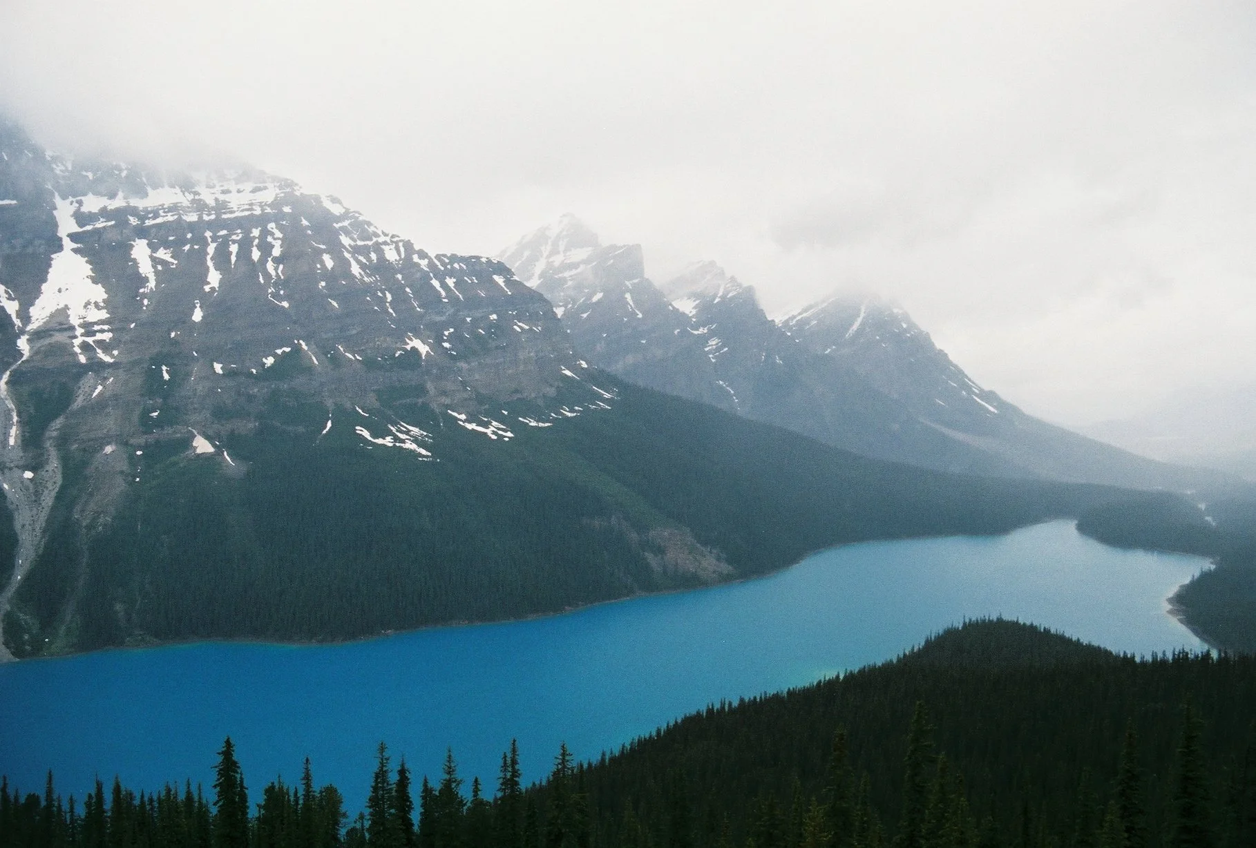 A mountain landscape with snow-capped peaks, a blue lake, and a forested foreground under a cloudy sky.