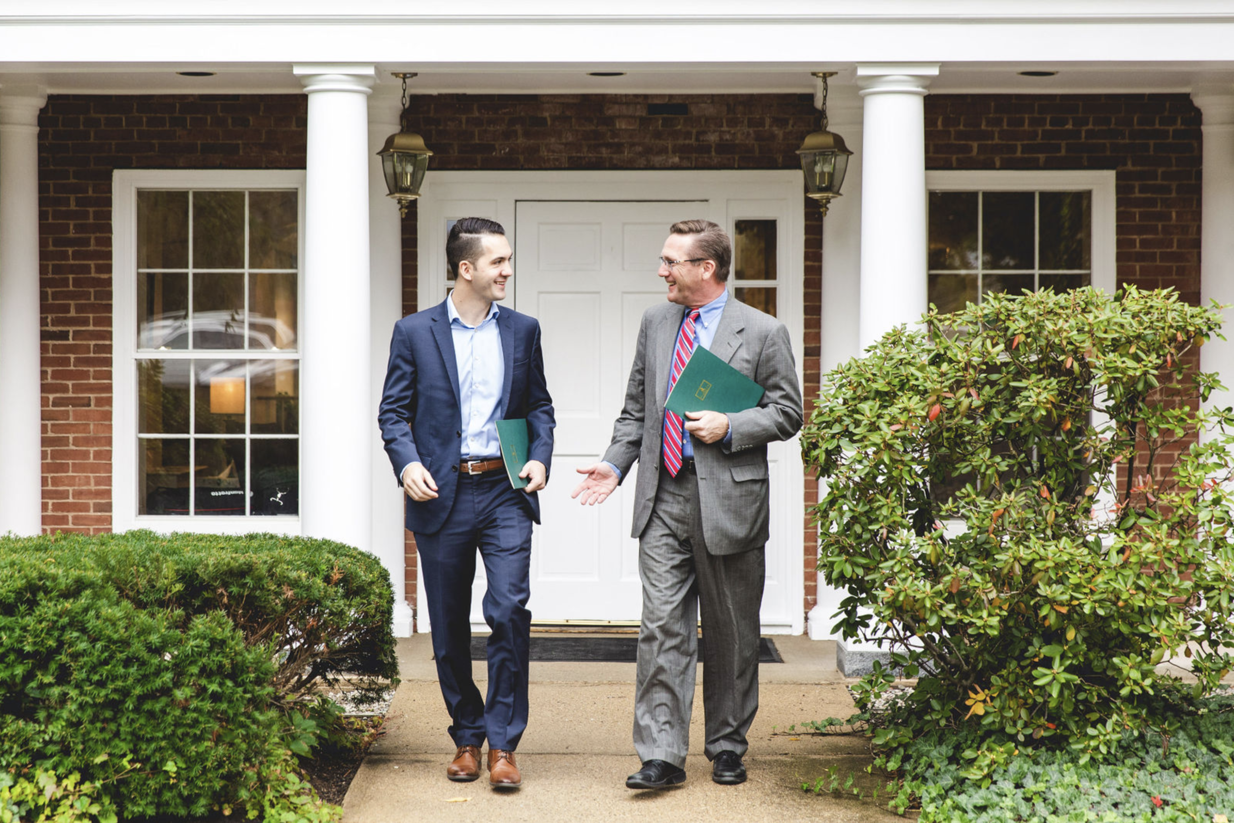 Two men in suits walking and talking on a front porch of a house, holding folders, and smiling.