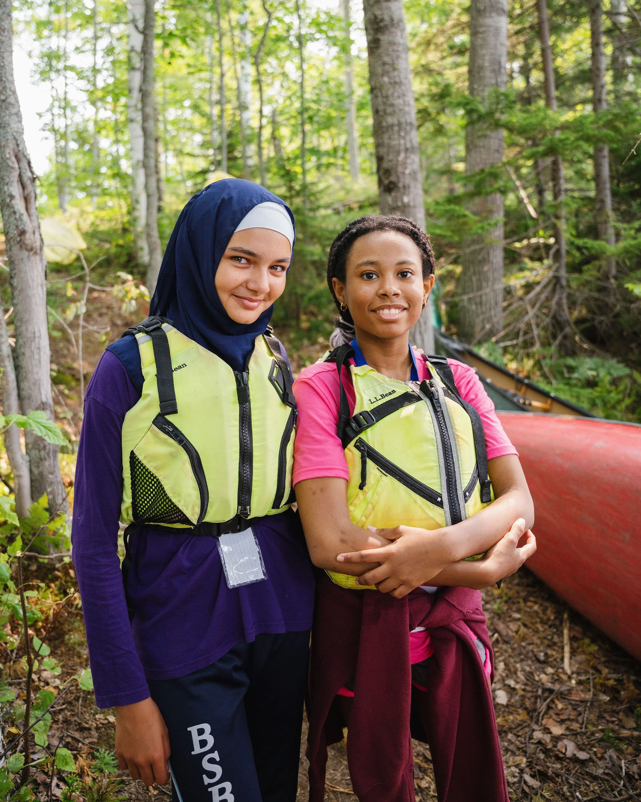 Two young women in hiking gear standing in a forest, smiling at the camera, with trees and a canoe in the background.