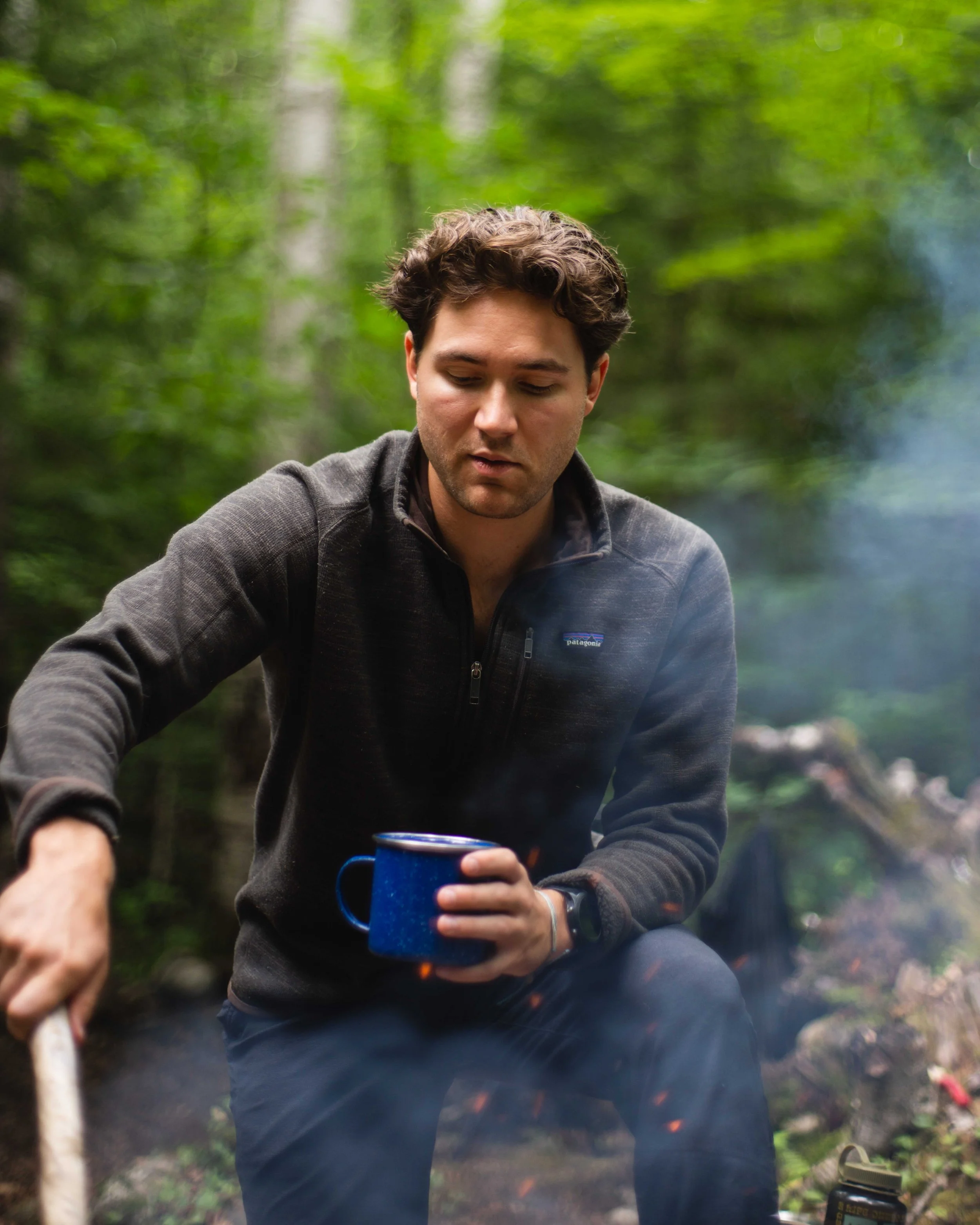 A young man with dark hair, wearing a black Patagonia fleece jacket, is sitting outdoors in a wooded area. He is holding a blue camping mug in his left hand and appears to be by a campfire, with smoke visible in the foreground. The background shows g