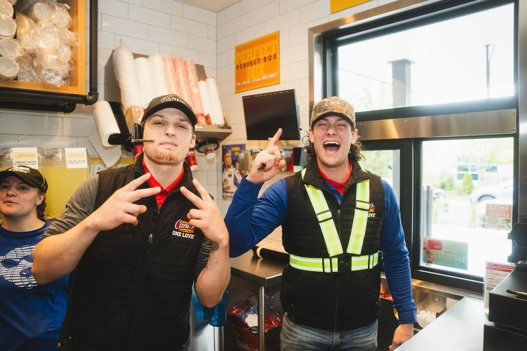 Two young men inside a fast food restaurant or pizza place, smiling and making gestures at the camera. One is wearing a black cap and vest, the other a camouflage cap and a reflective vest. There is a woman in the background and store shelves with cu