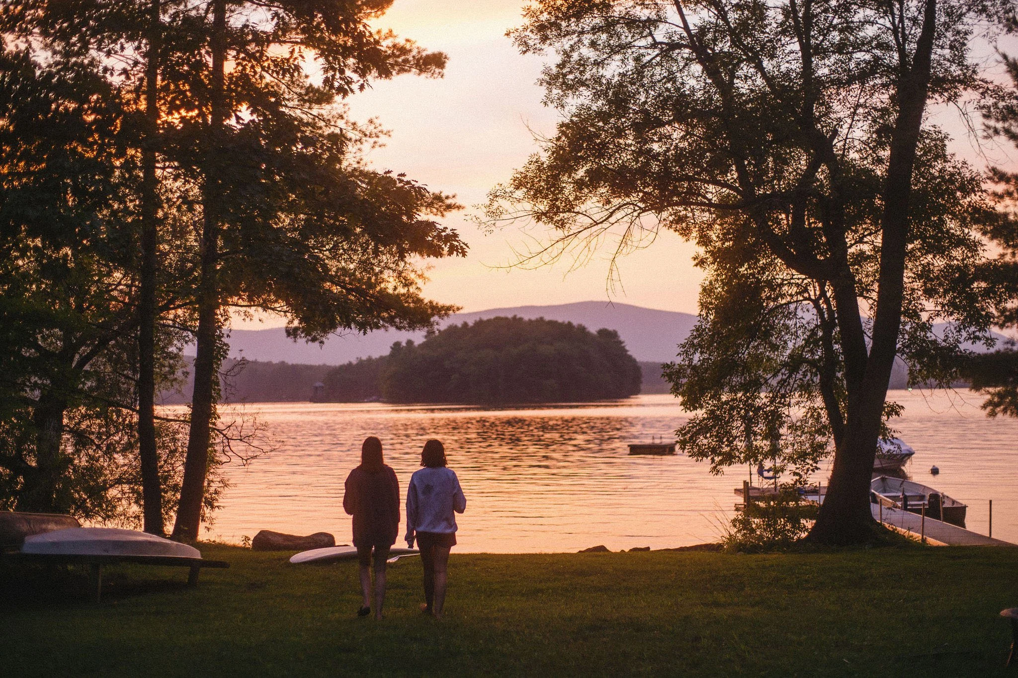 Two women walk along the grassy shore of a lake at sunset, framed by tall trees, with boats docked at a pier and mountains in the background.