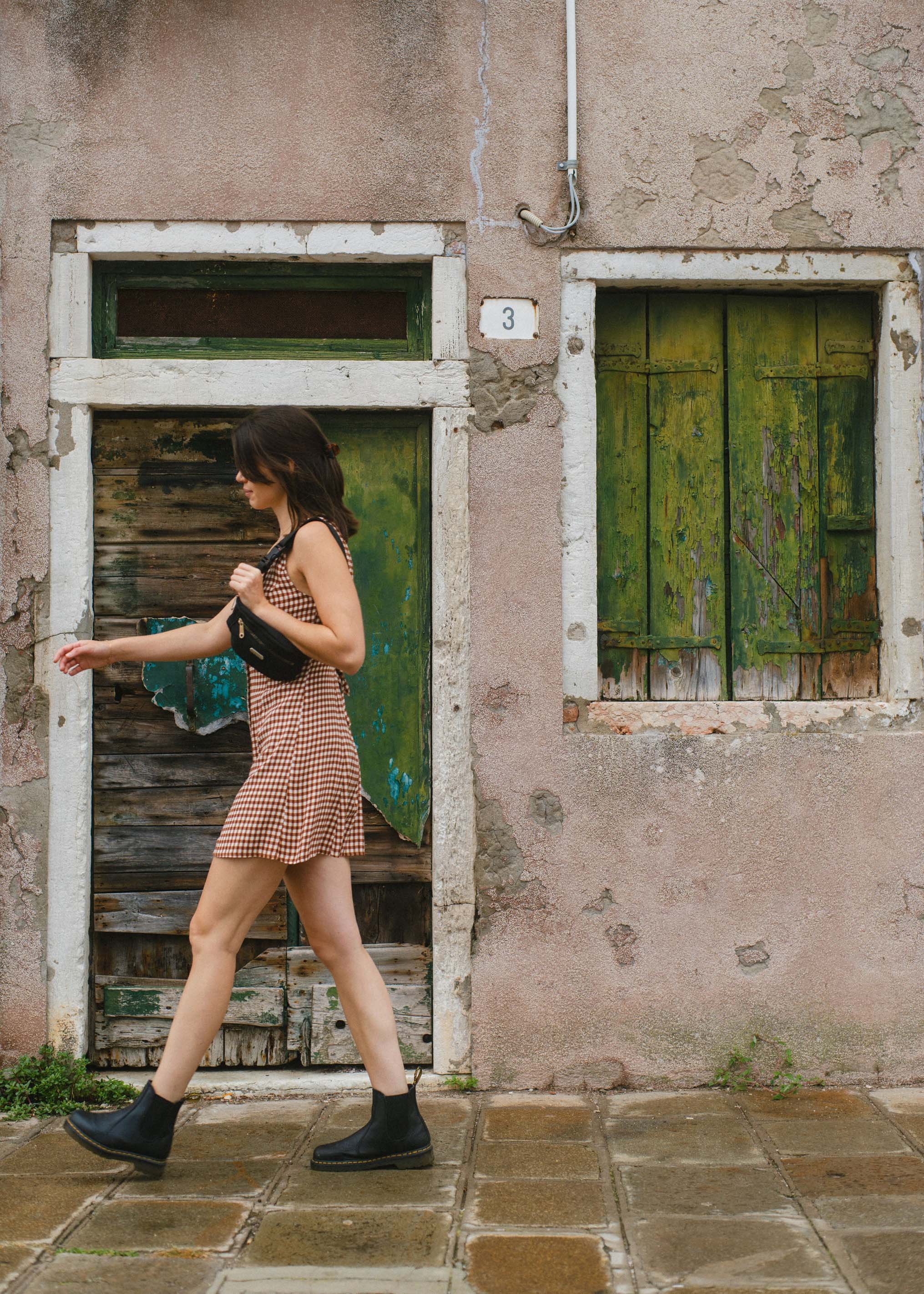 A woman in a checkered dress and black boots walking past a weathered building with peeling paint and green wooden shutters.