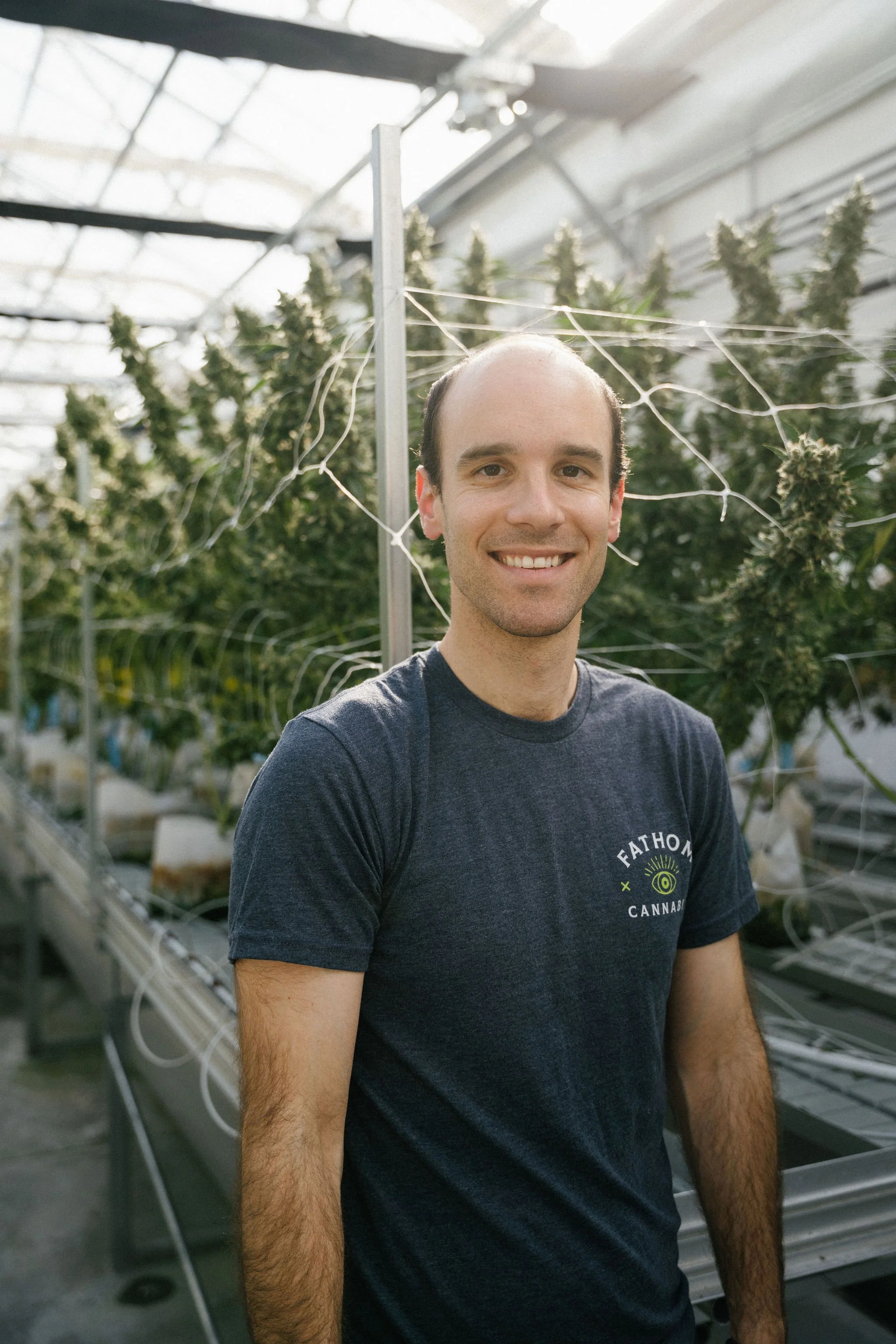 A man smiling in a greenhouse surrounded by cannabis plants with flowering buds.