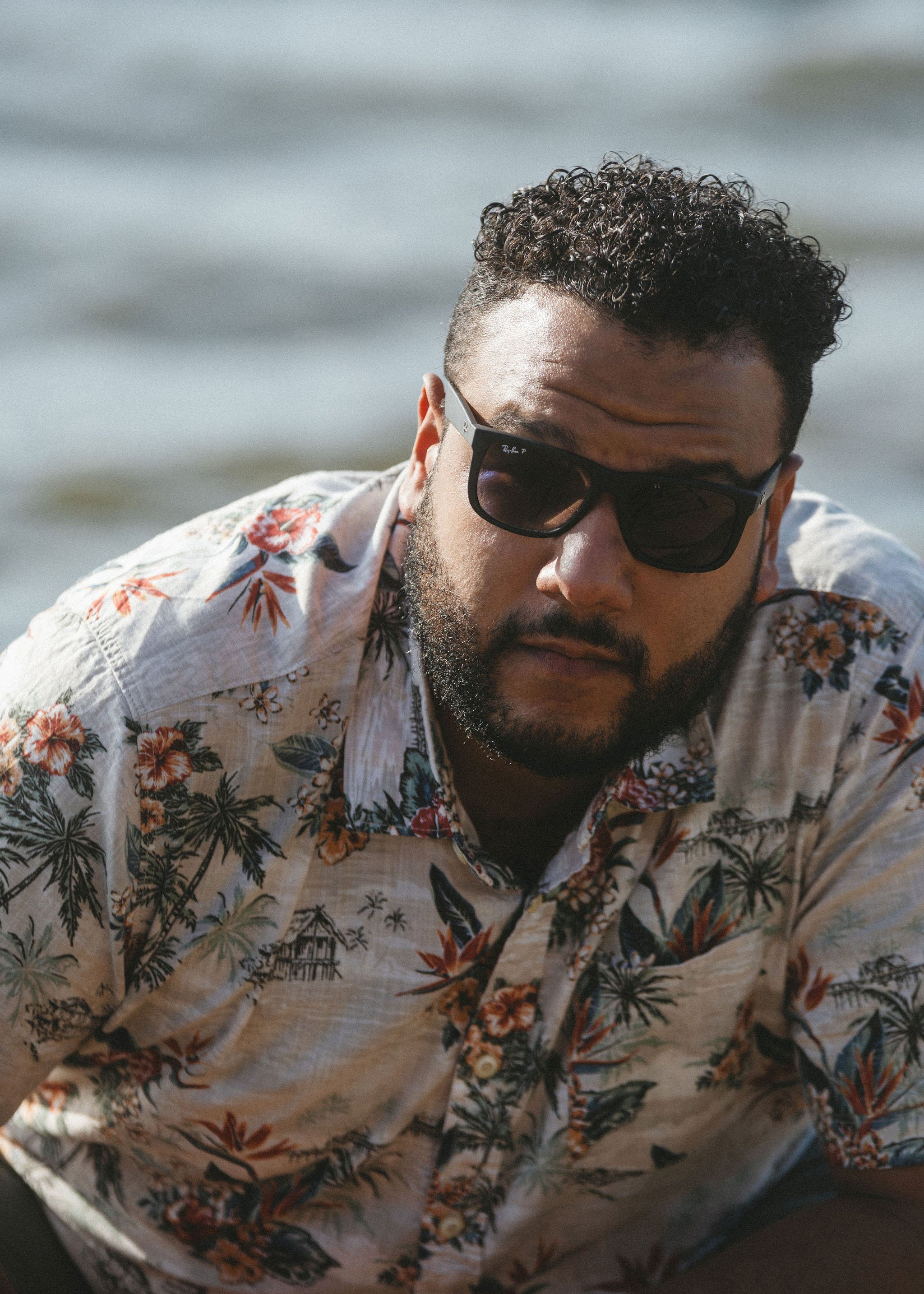 A man with curly hair and a beard wearing sunglasses and a floral Hawaiian shirt, leaning forward outdoors near water.