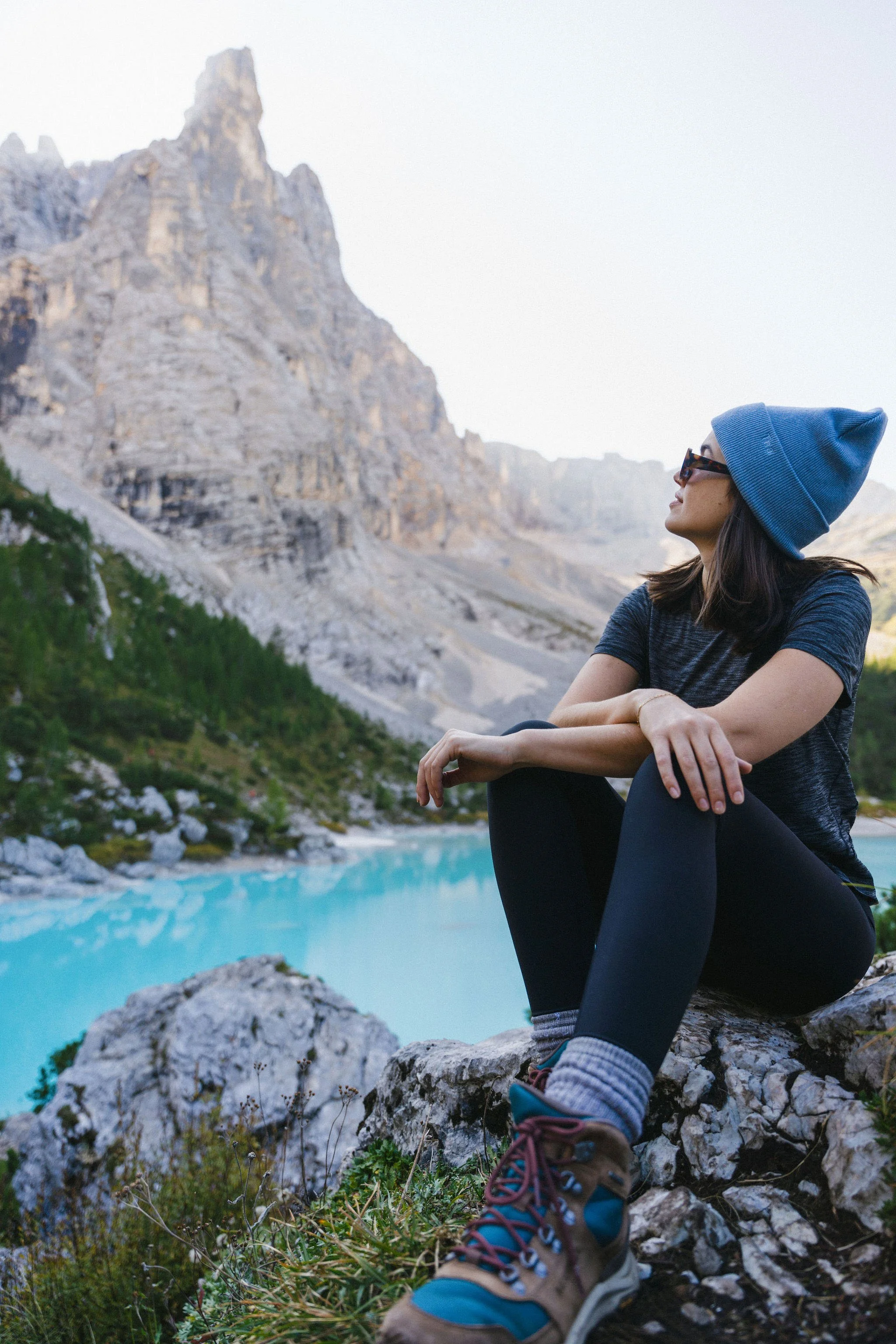 Woman sitting on rocks by a bright blue lake in a mountainous landscape, wearing a blue beanie, sunglasses, and hiking boots.