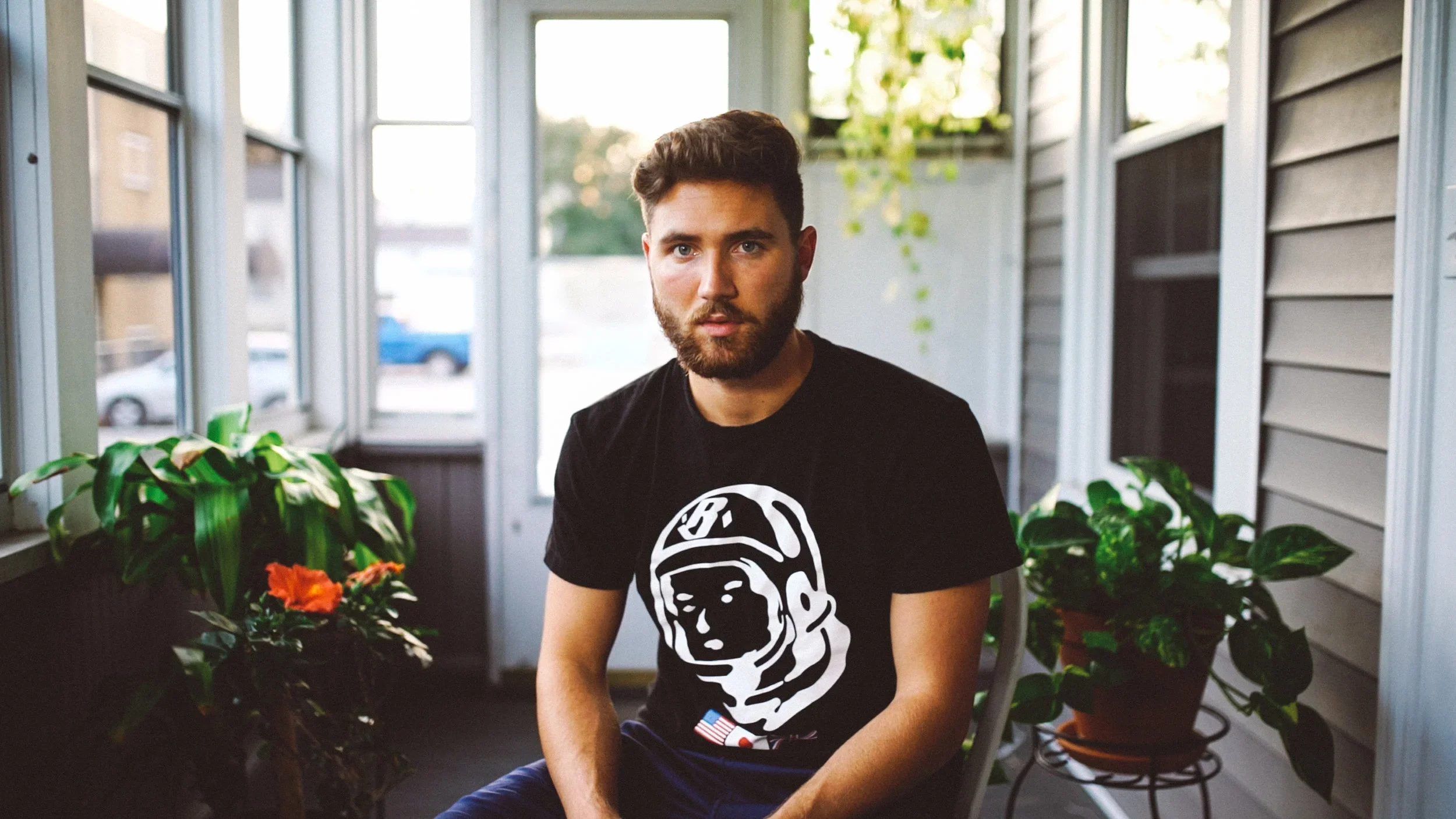 A young man with a beard and short brown hair sitting in a porch area with plants, wearing a black T-shirt with a white astronaut face design.