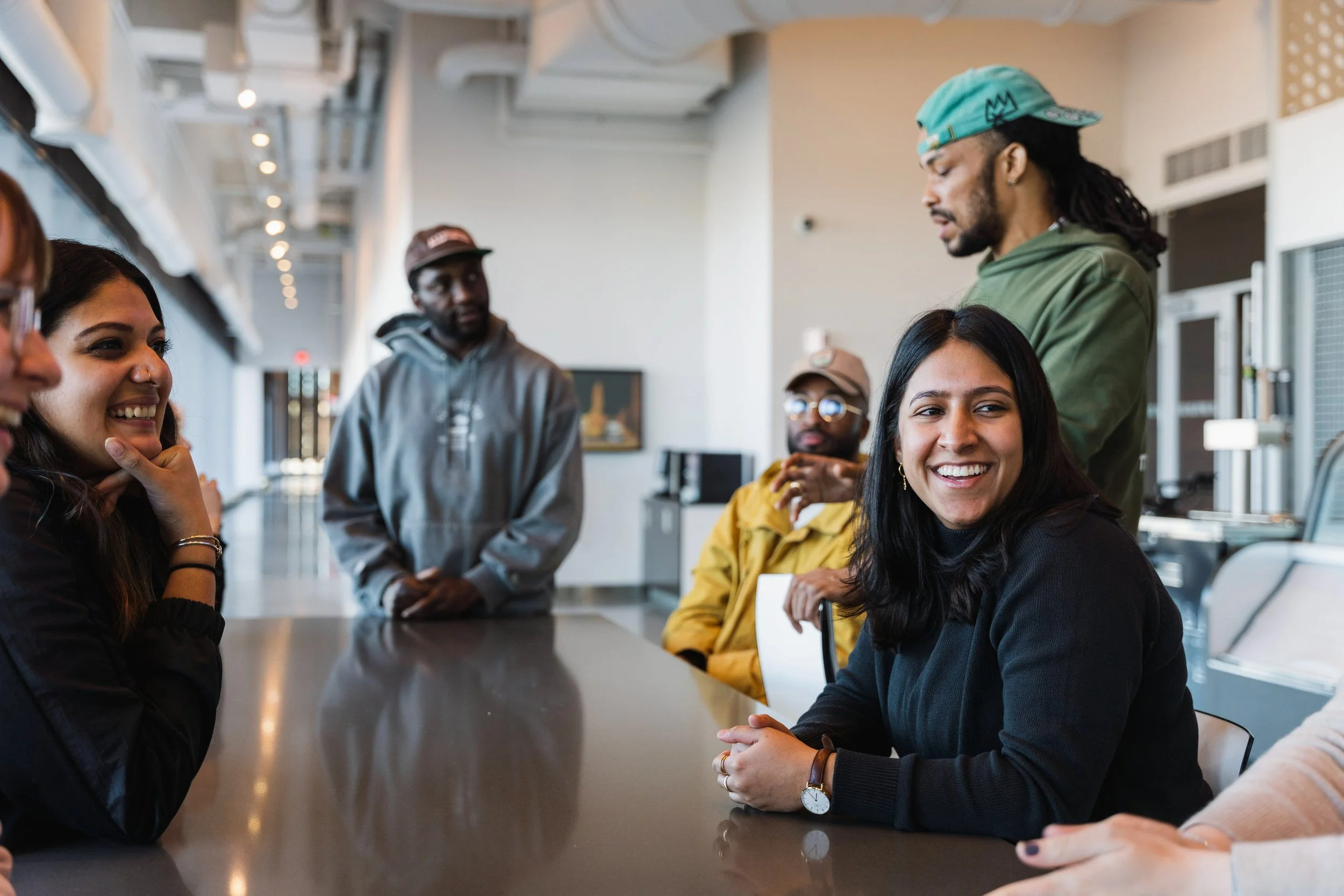 Group of diverse young adults sitting and standing around a table in a modern office or cafe, engaging in conversation and smiling.